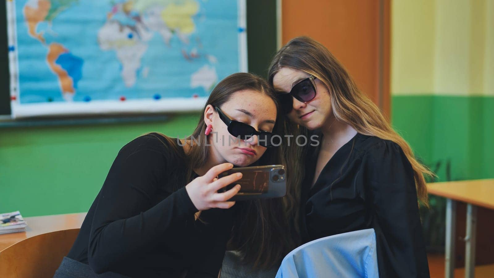 Two female high school students wearing sunglasses are using a smartphone in a classroom, potentially distracting themselves from their studies