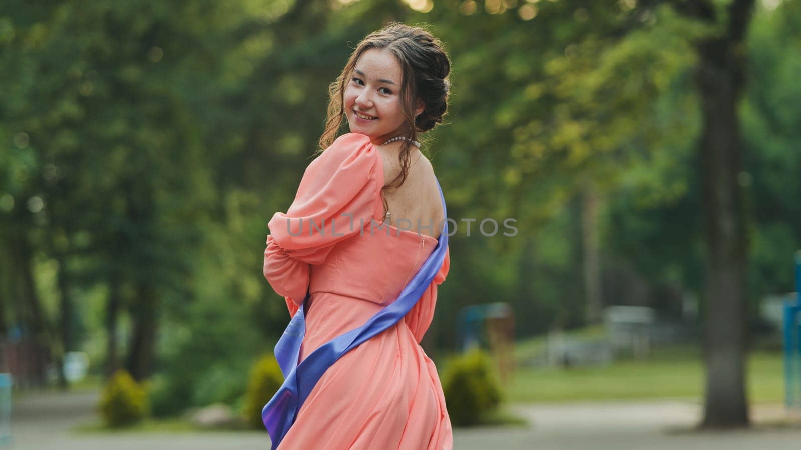 Female graduate beaming, wearing formal attire with academic stole, spinning gracefully within green campus landscape