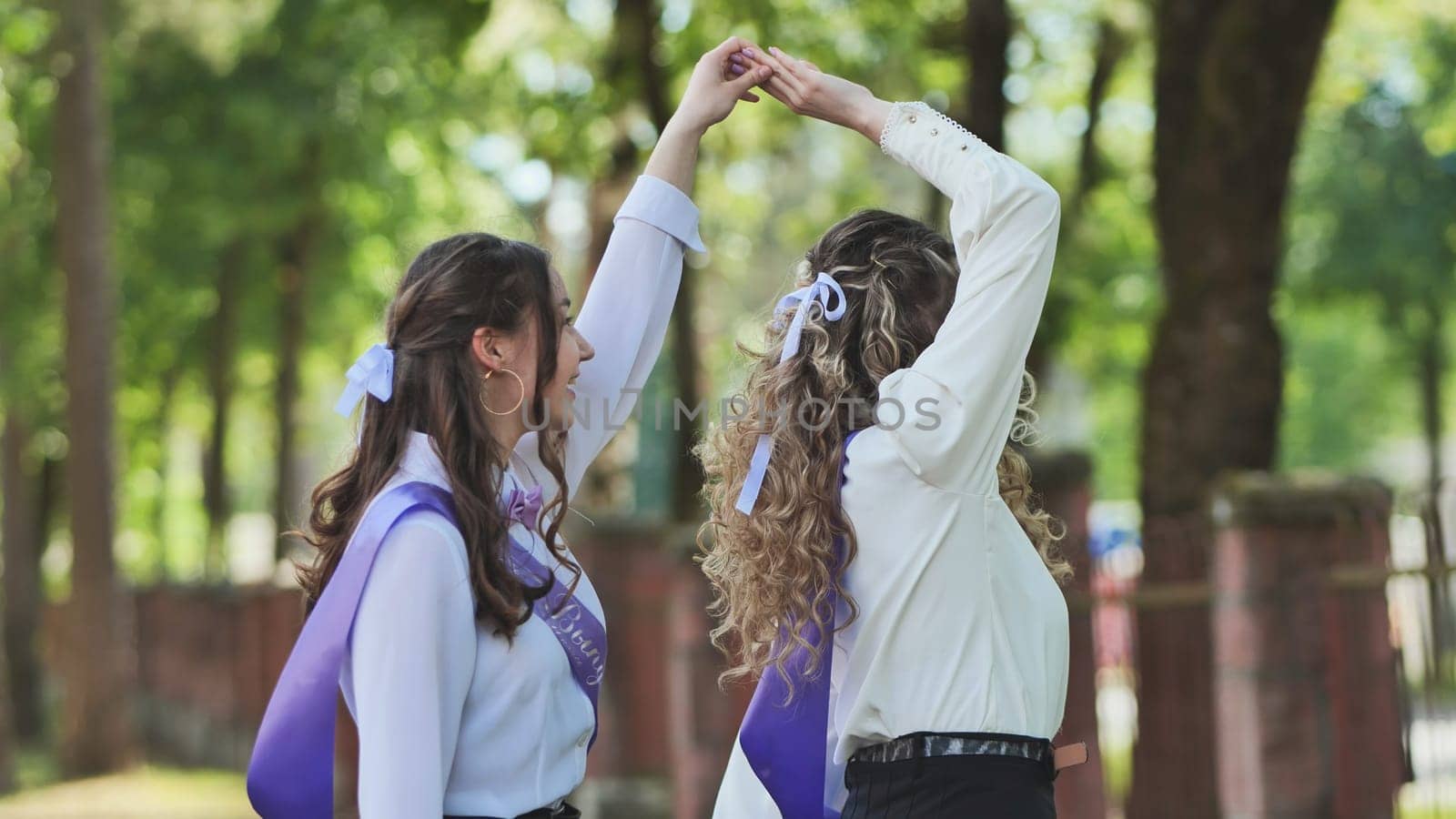 Two happy graduates are holding hands and dancing together, celebrating their last school day in a green park
