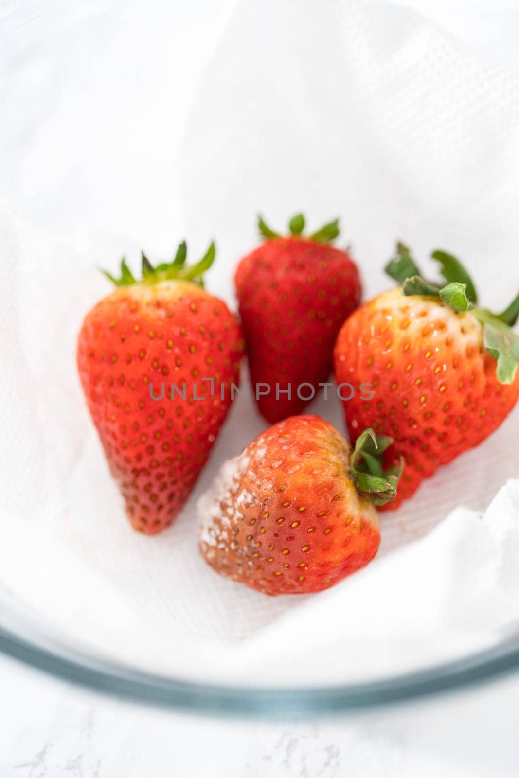 Bright red strawberries, interspersed with signs of mold, rest in a glass bowl lined with a paper towel on a white napkin, indicating improper storage techniques.