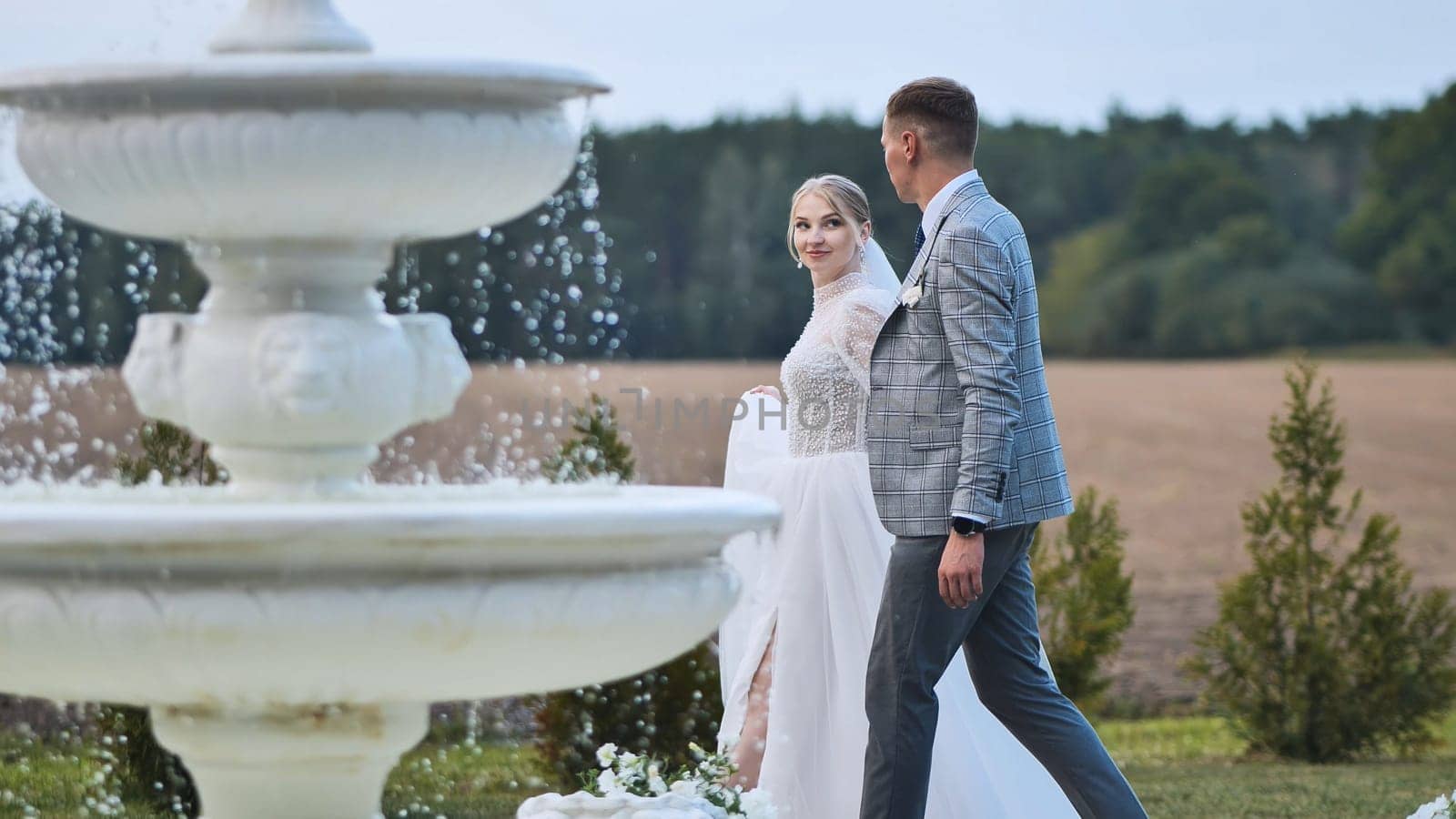 Newlywed couple holding hands beside elegant white fountain, sharing intimate moment amid lush park greenery during wedding celebration