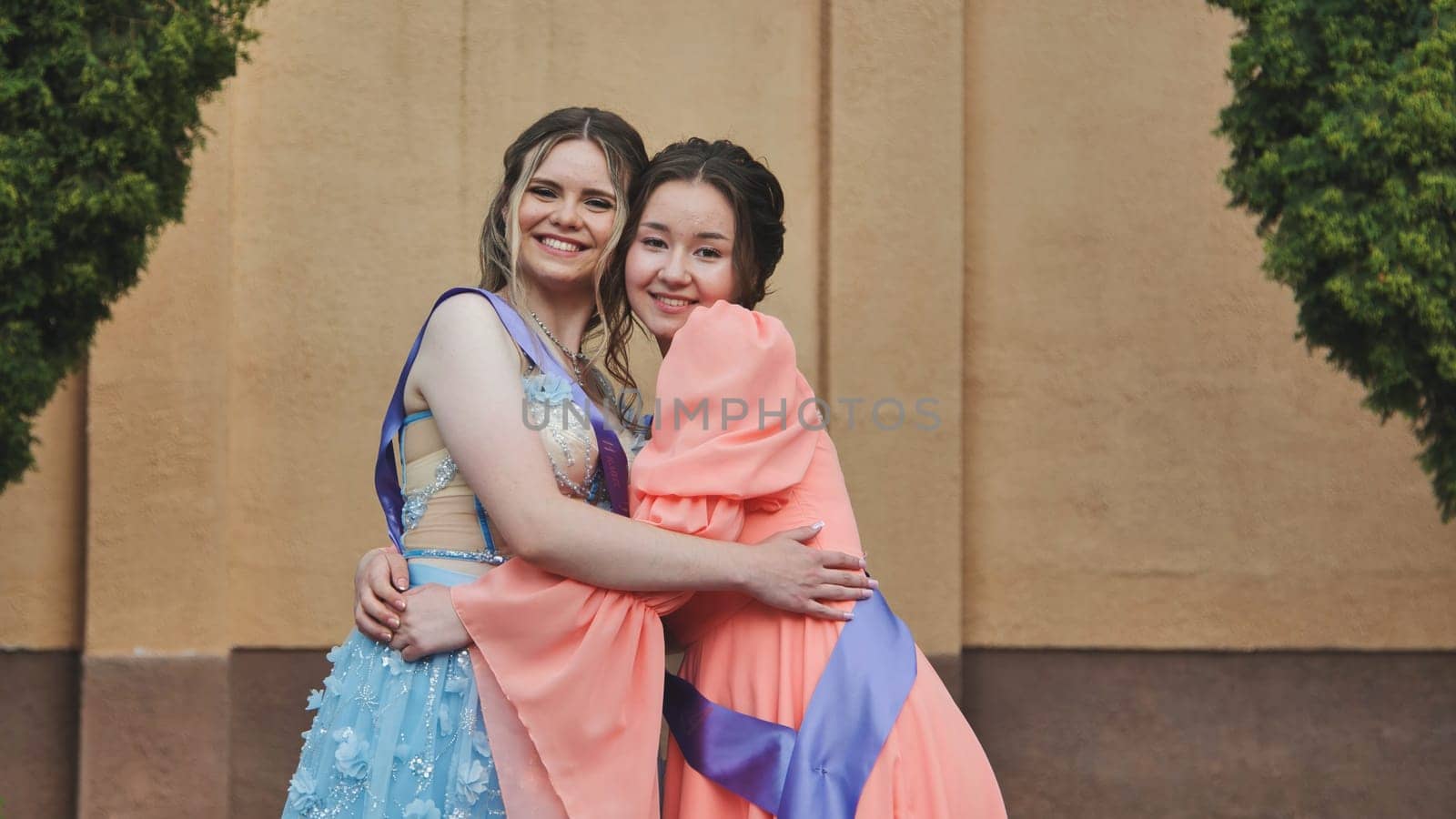Two happy graduates in elegant dresses are hugging each other, celebrating their academic achievement and friendship