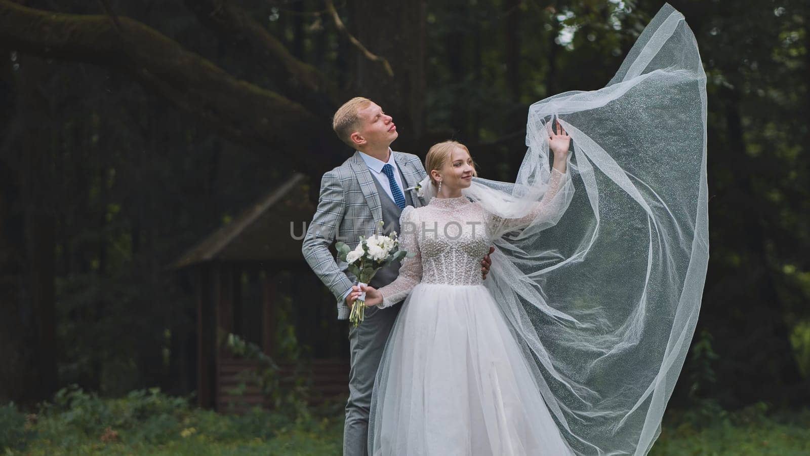 Newlyweds holding a bouquet and raising a long veil in a windy forest, celebrating their wedding day with joy and love