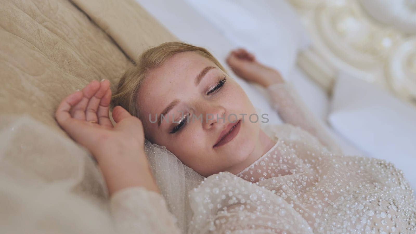 Beautiful blond bride resting on a bed wearing a white wedding dress with pearl decorations and veil, enjoying a moment of peace on her wedding day