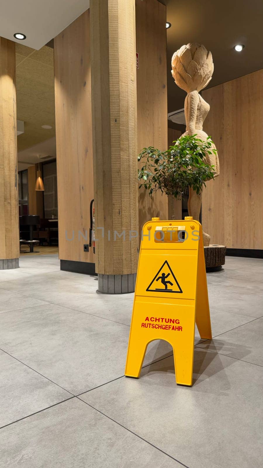 25.03.2025 Germany: Caution Wet Floor Sign in Modern Wooden Interior with Decor in McDonalds. Bright yellow caution wet floor sign displayed on tiled floor in a cozy wooden interior with decor.