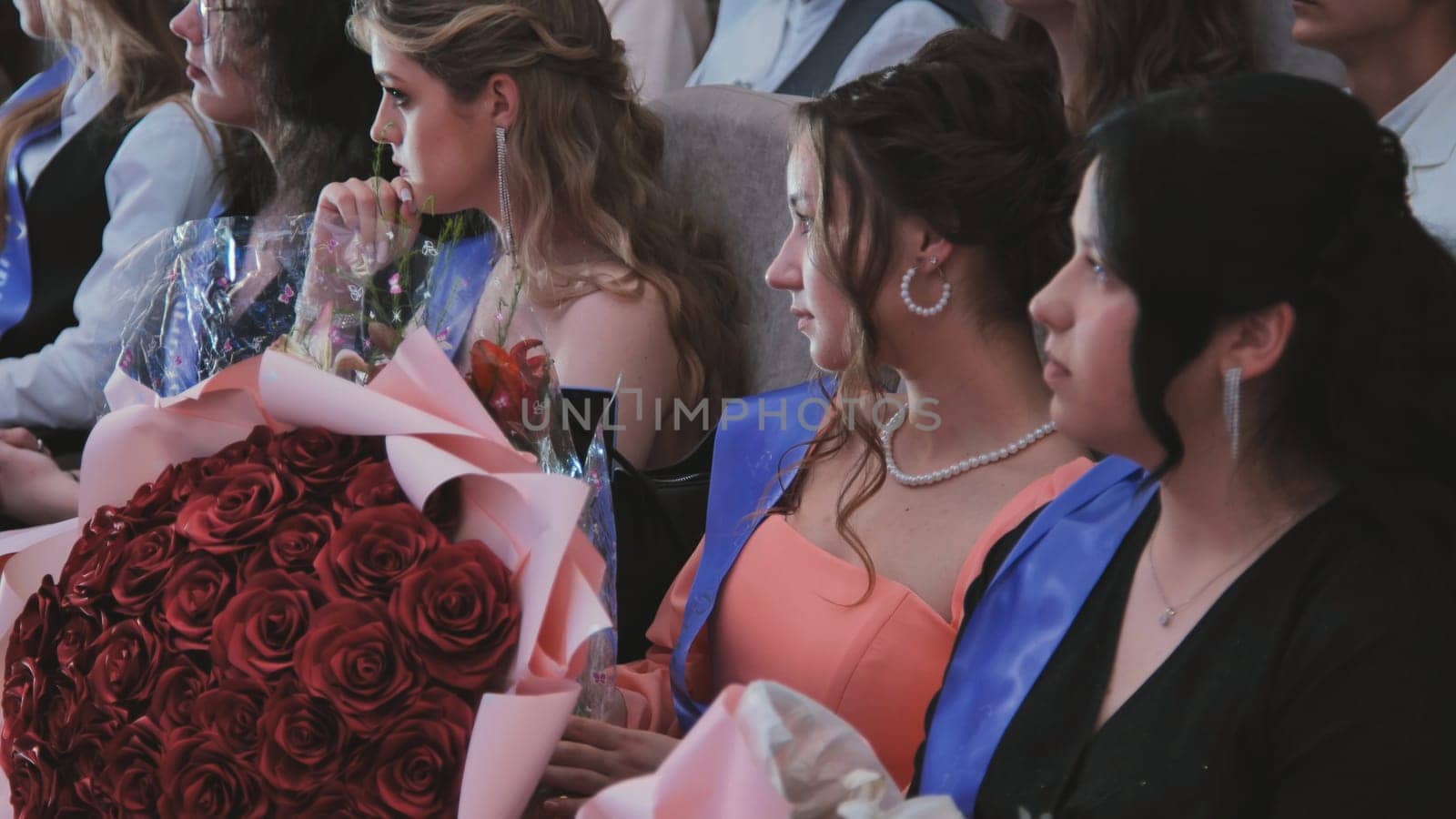 Female graduates attentively listening during graduation ceremony, clutching bouquets of vibrant red roses