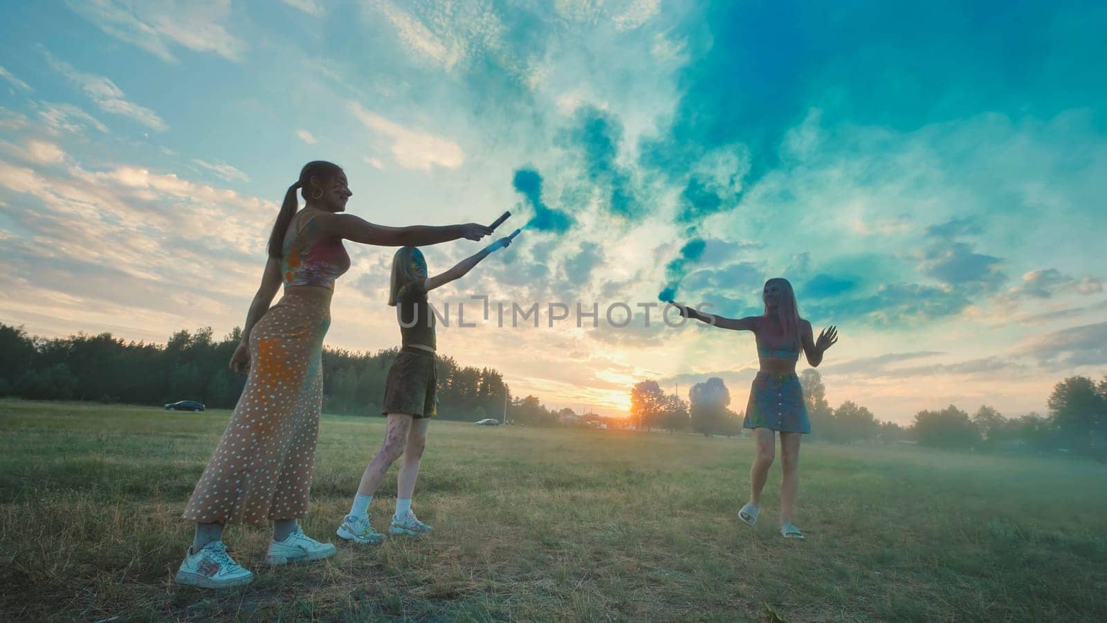 Three girls celebrating their friendship by playing with colored smoke bombs in a field at sunset
