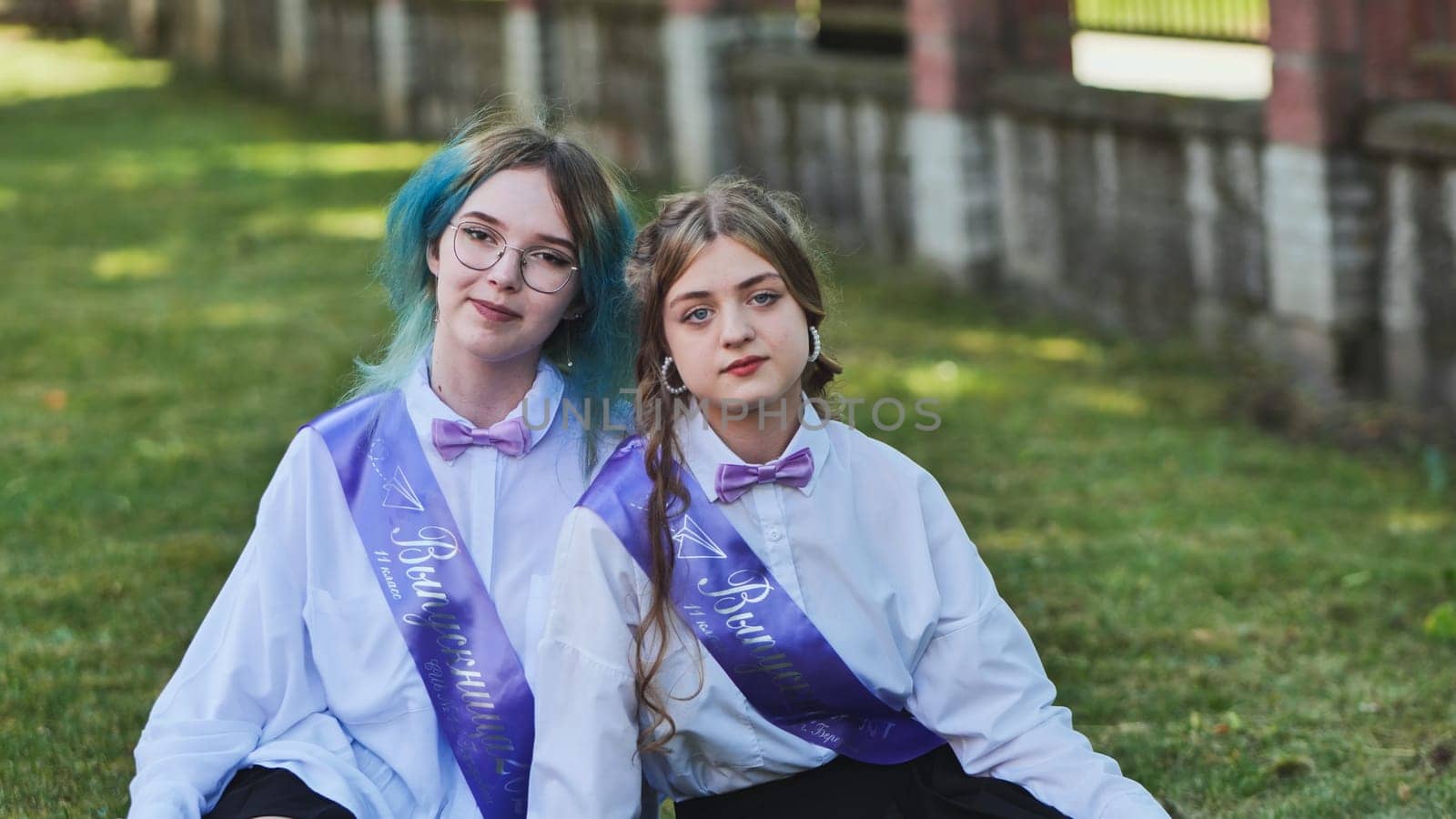 Two joyful graduates enjoying their final school day, sitting on the grass in the schoolyard, donning sashes and savoring the moment as best friends and classmates