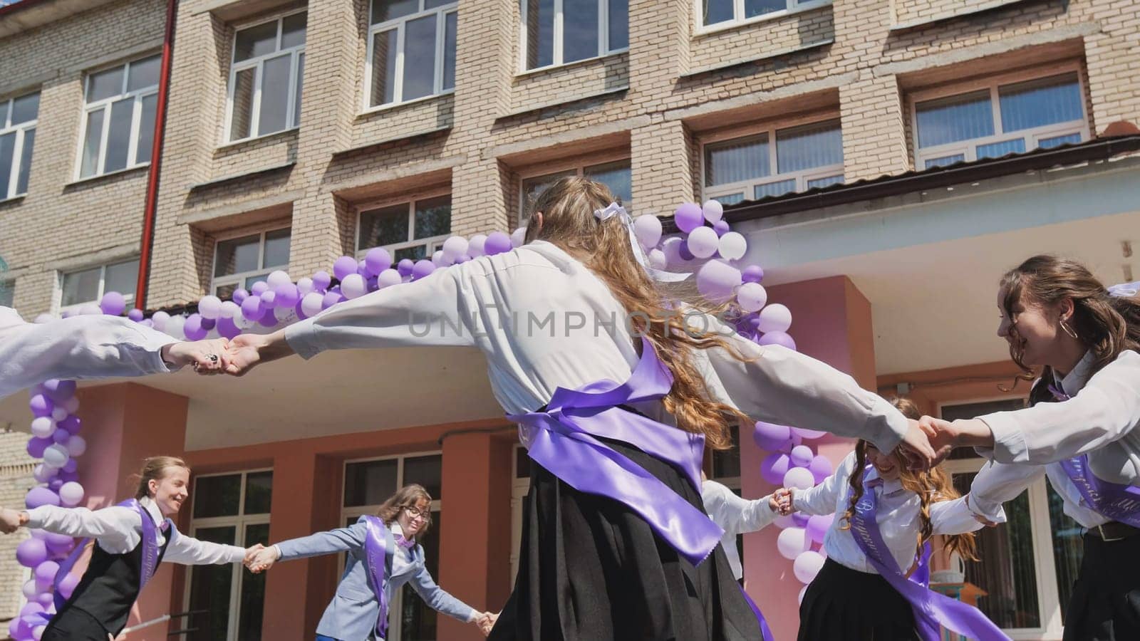 Group of happy students dancing together in a circle, celebrating their last school day with purple and white balloons and ribbons