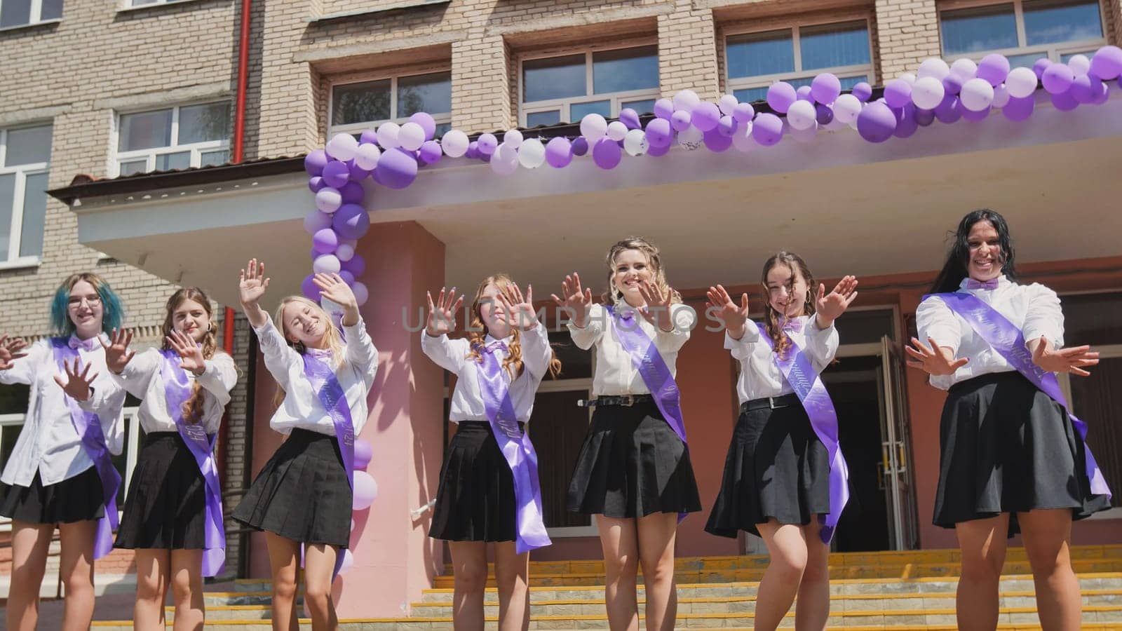 Group of cheerful female graduates waving goodbye on their last school day, celebrating the end of academic year with purple ribbons and balloons