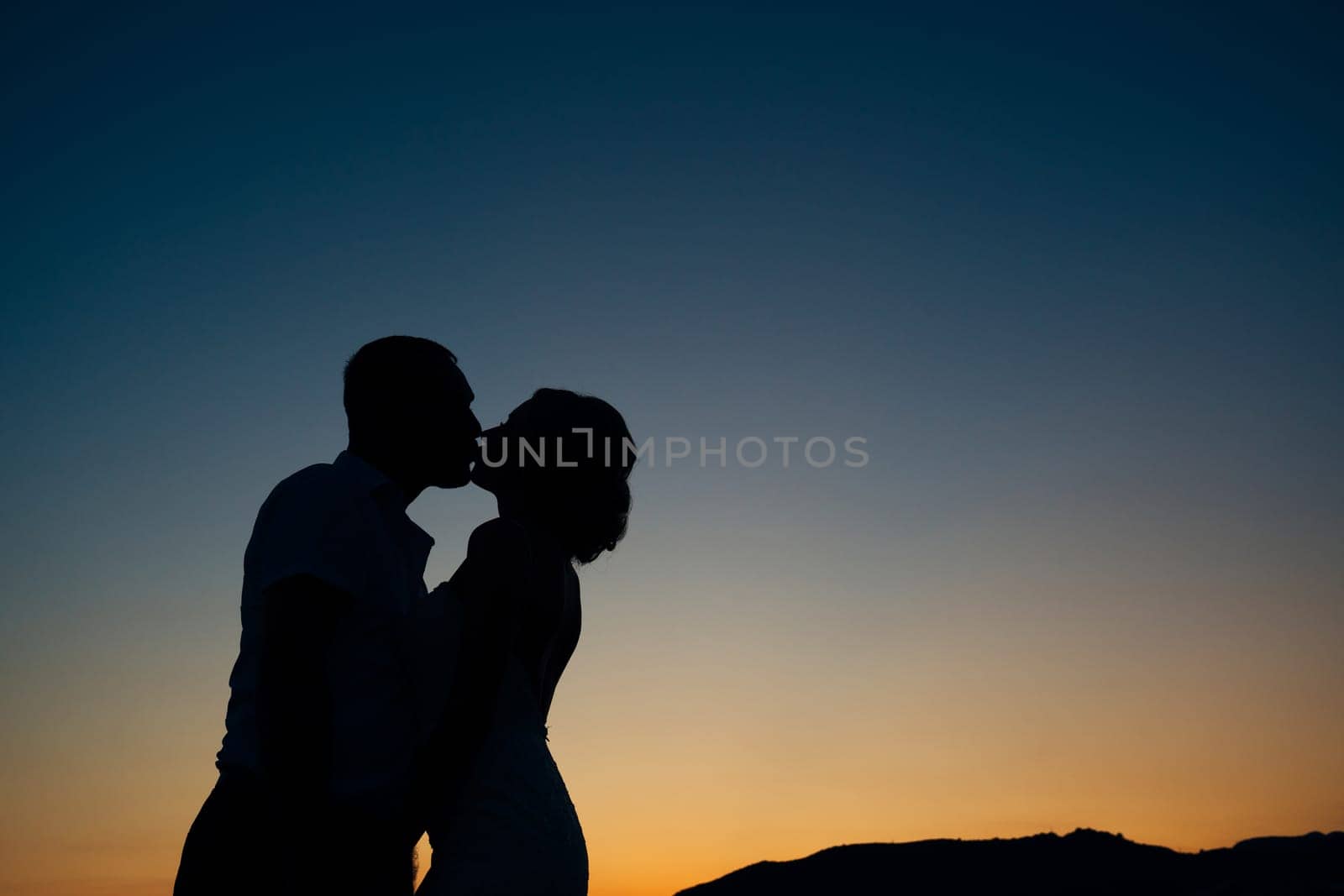 Silhouettes of kissing bride and groom against the backdrop of a sunset over the mountains. High quality photo