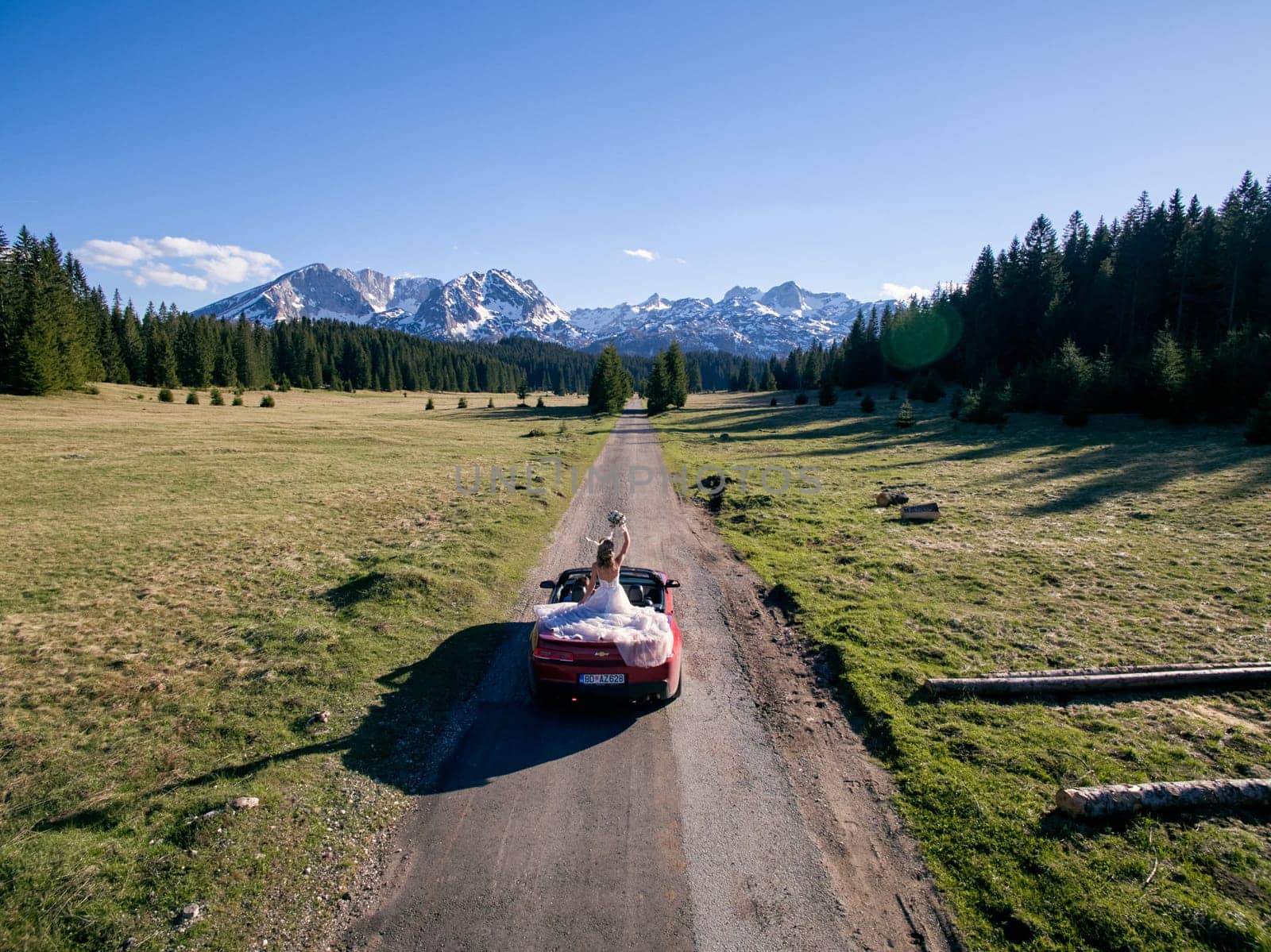 Bride with a bouquet stands in a cabriolet driving along the road in a mountain valley. Back view. Drone. High quality photo