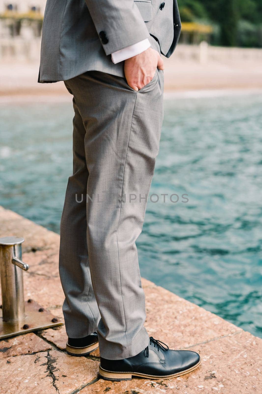 Man stands with his hands in his trouser pockets on the pier near the bollard. Cropped. Faceless. High quality photo