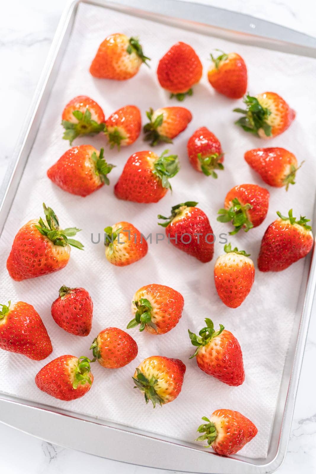 Juicy red strawberries, freshly washed, are spread out to dry on a baking sheet, carefully lined with paper towels to absorb excess moisture and prevent mold.