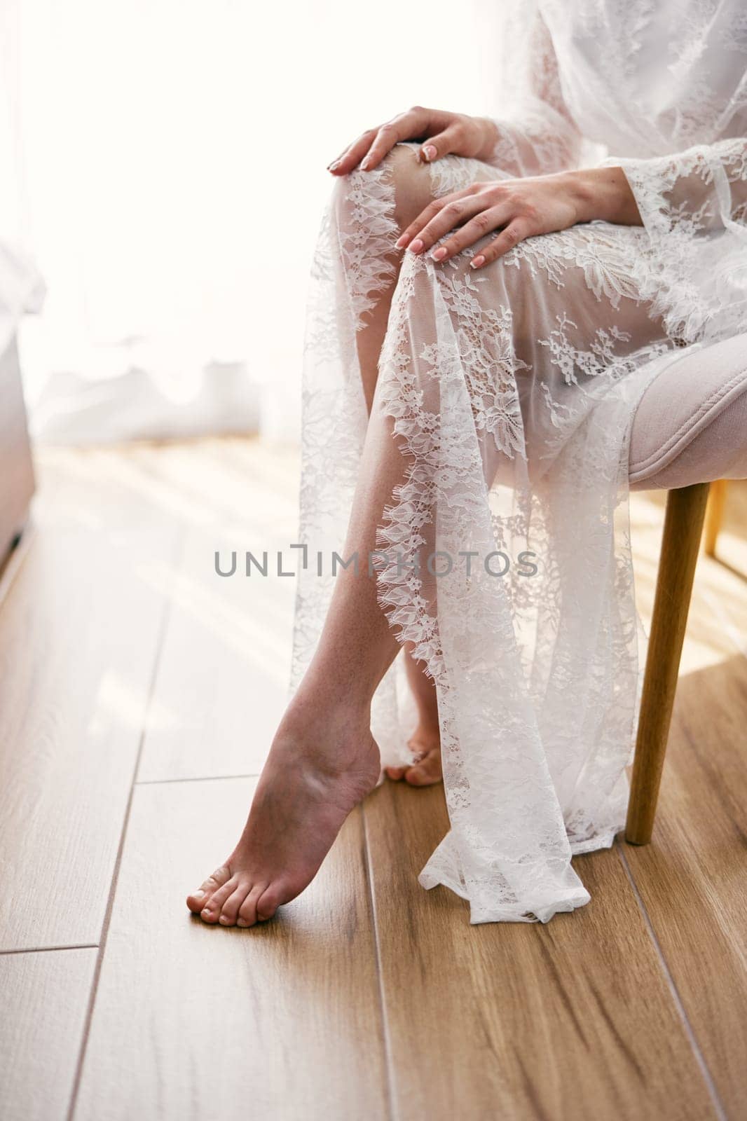 Bride in a lace robe sits barefoot on an armchair with her hands on her knees. Cropped. Faceless. High quality photo