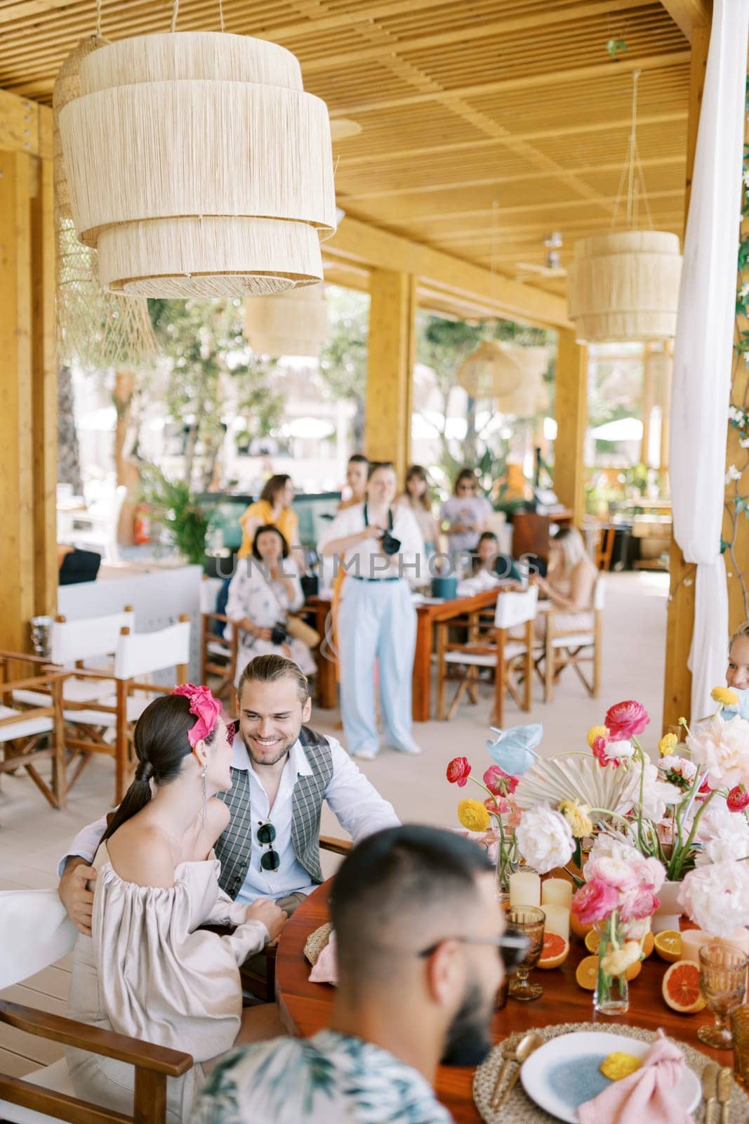 Bride and groom look at each other while sitting on chairs at a festive table with guests on the terrace. High quality photo