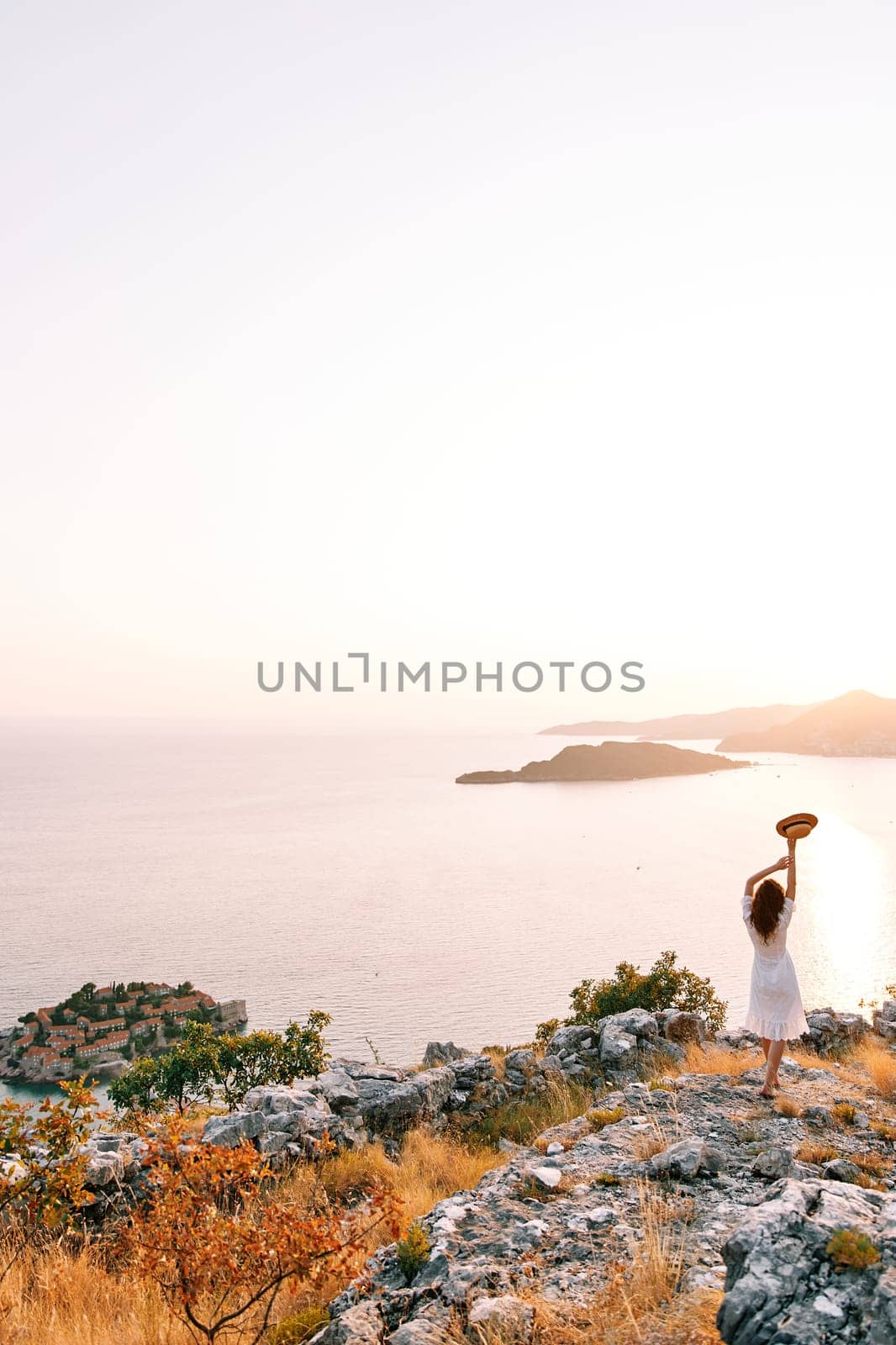 Girl with a hat in her hands above her head stands on a mountain above the island of Sveti Stefan. Montenegro. Back view. High quality photo