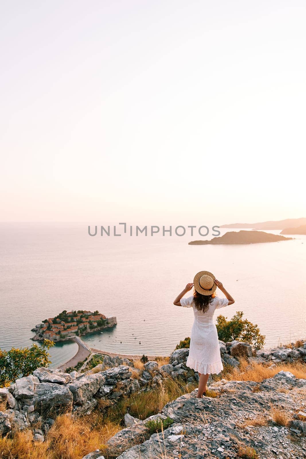 Girl straightens her hat standing on the mountain and looks at the island of Sveti Stefan. Montenegro. Back view. High quality photo