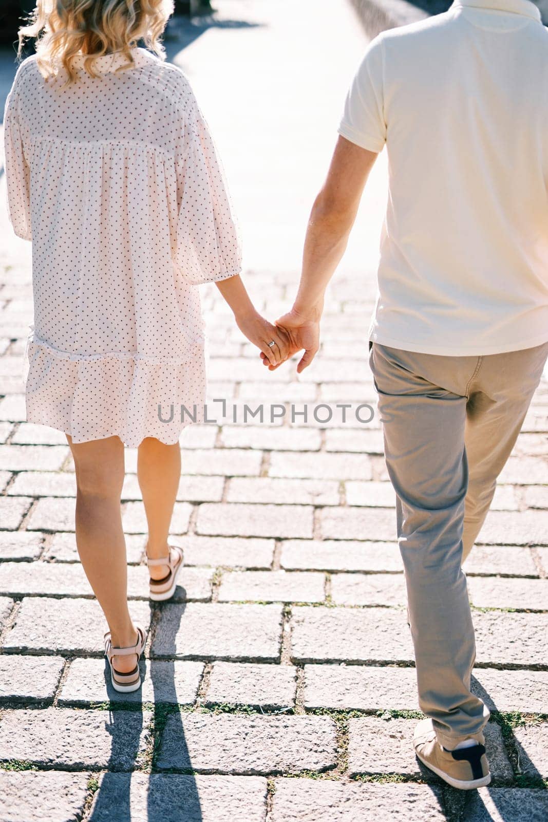 Man and woman walk holding hands along a sunny paved road. Back view. Cropped. High quality photo