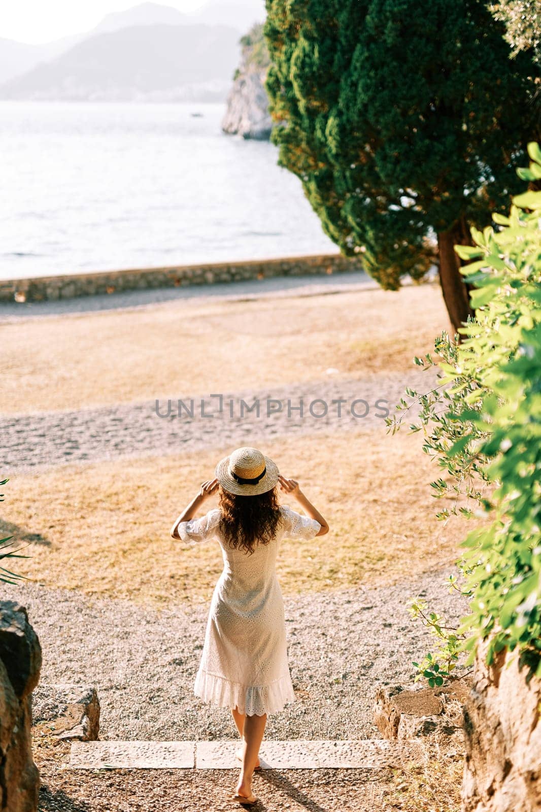 Young woman in a sundress goes to the beach, holding her hat on her head. Back view. High quality photo