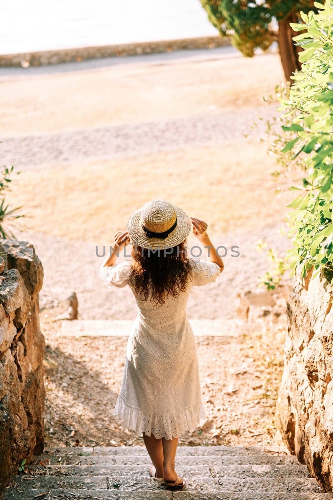 Girl goes down the stone steps to the beach, touching the hat on the head. Back view. High quality photo