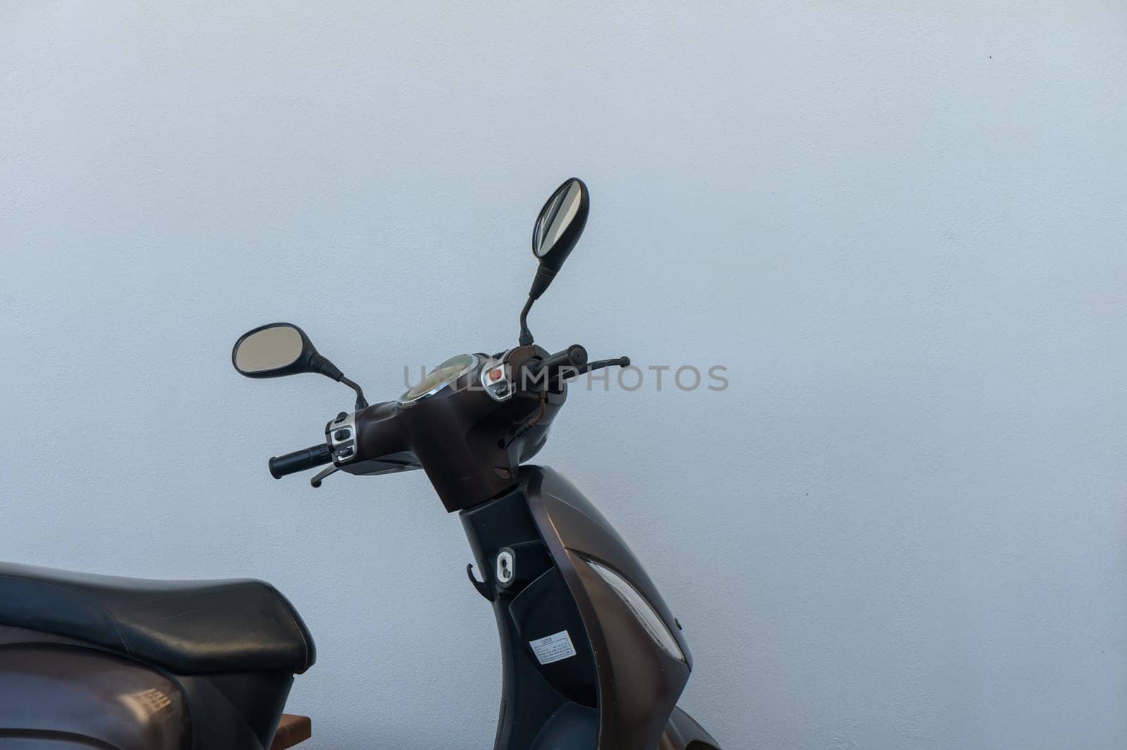 A sleek, brown motorcycle stands idle beside a simple white wall, symbolizing urban mobility and freedom. The shiny mirrors reflect a tranquil moment in a bustling city environment.