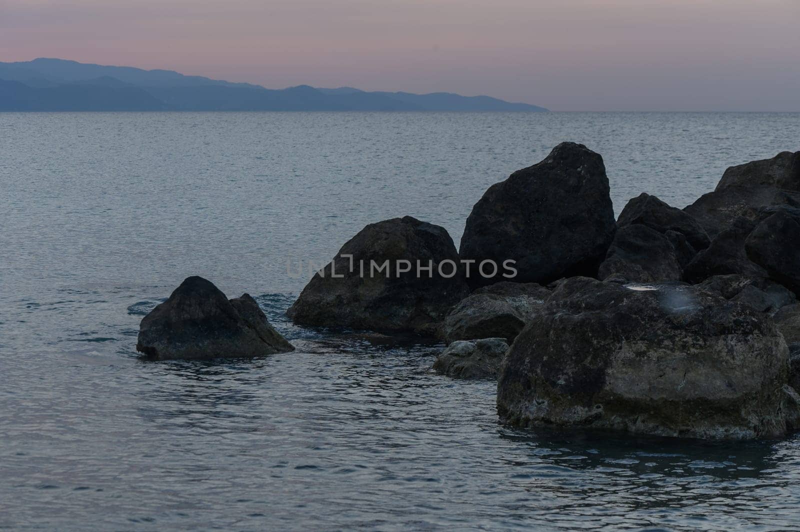 As dusk settles over the coastline, rugged rocks emerge from calm waters, reflecting soft hues of twilight and inviting peaceful contemplation by the shore.