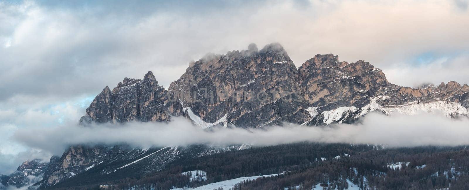 Aerial view of rugged Dolomite peaks near Cortina d'Ampezzo, Italy, with snow covered slopes, low clouds, and visible ski trails and structures.