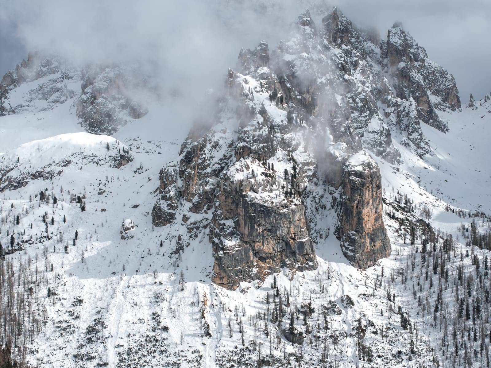 A dramatic snow covered mountain in the Dolomites near Cortina, Italy, with rugged peaks, sparse trees, and mist partially shrouding the cliffs.