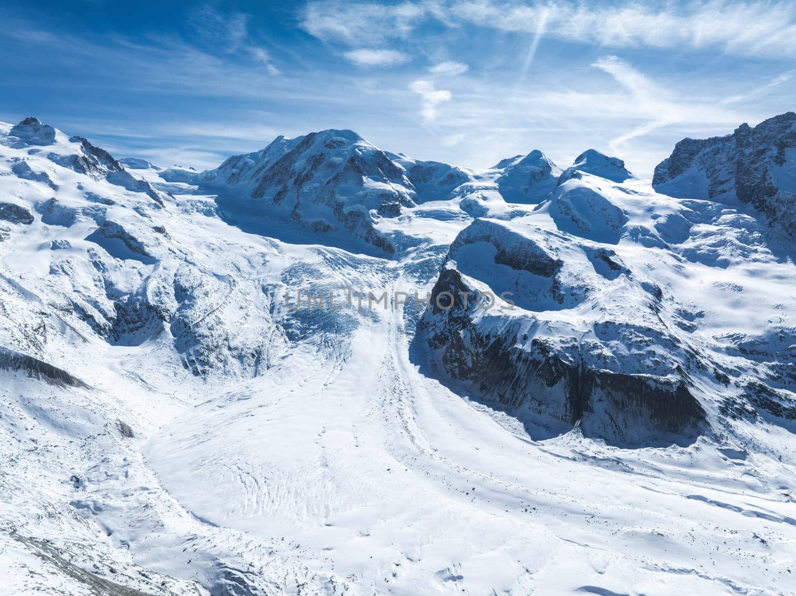 Aerial view of a glacier flowing through a valley in the Alps, surrounded by towering snow covered peaks under a bright blue sky with scattered clouds.