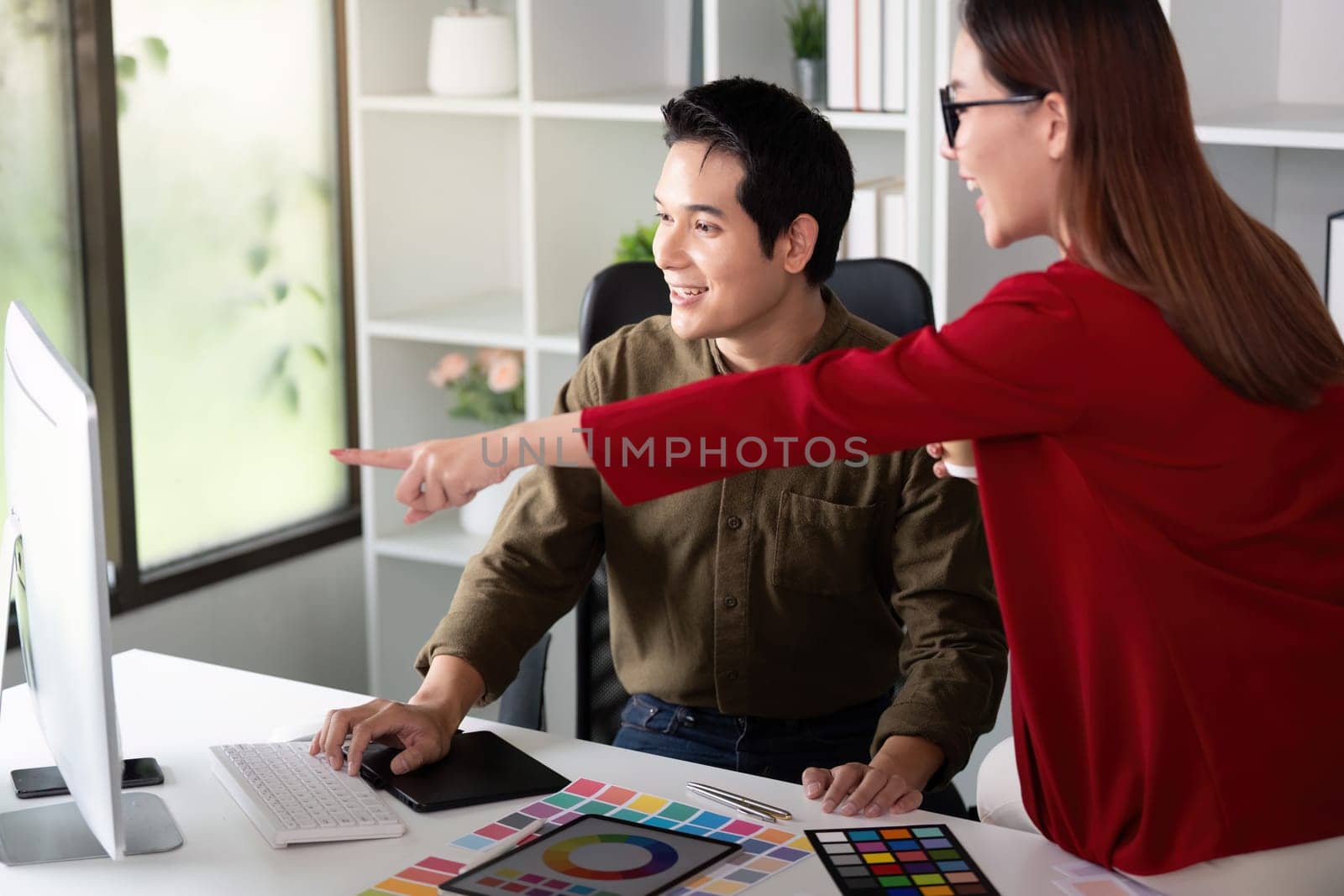 In a bright office, a female designer points out color choices while her colleague reviews digital designs, emphasizing collaboration in a colorful workspace.