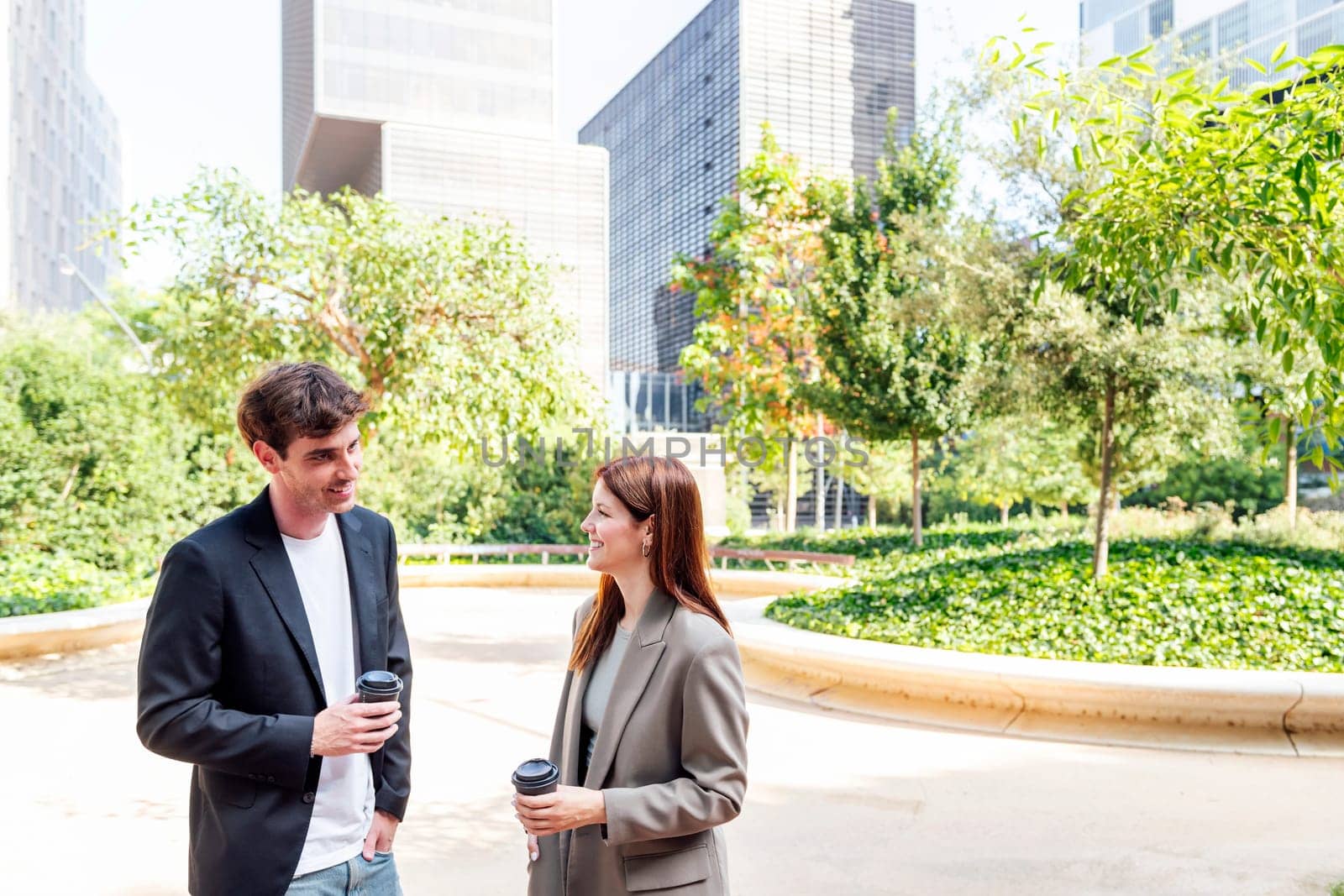 smiling man and a woman standing in a park near of office buildings talking while taking coffee, they are smiling and enjoying with the company, concept of business, copyspace for text
