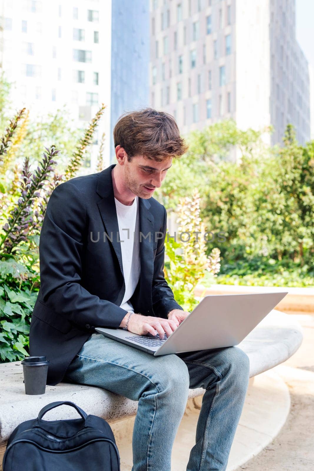 smiling young man using a laptop sitting on a bench of a park near of office buildings, concept of business and technology for remote work