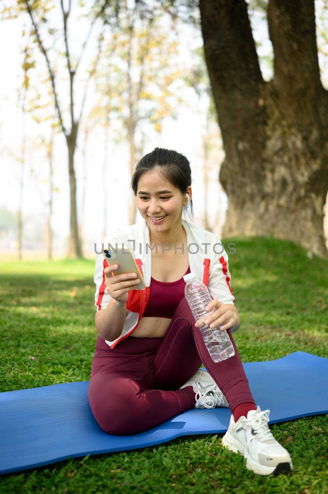 Cheerful woman relaxing after workout and using mobile phone, sitting on mat under trees in a sunny park.