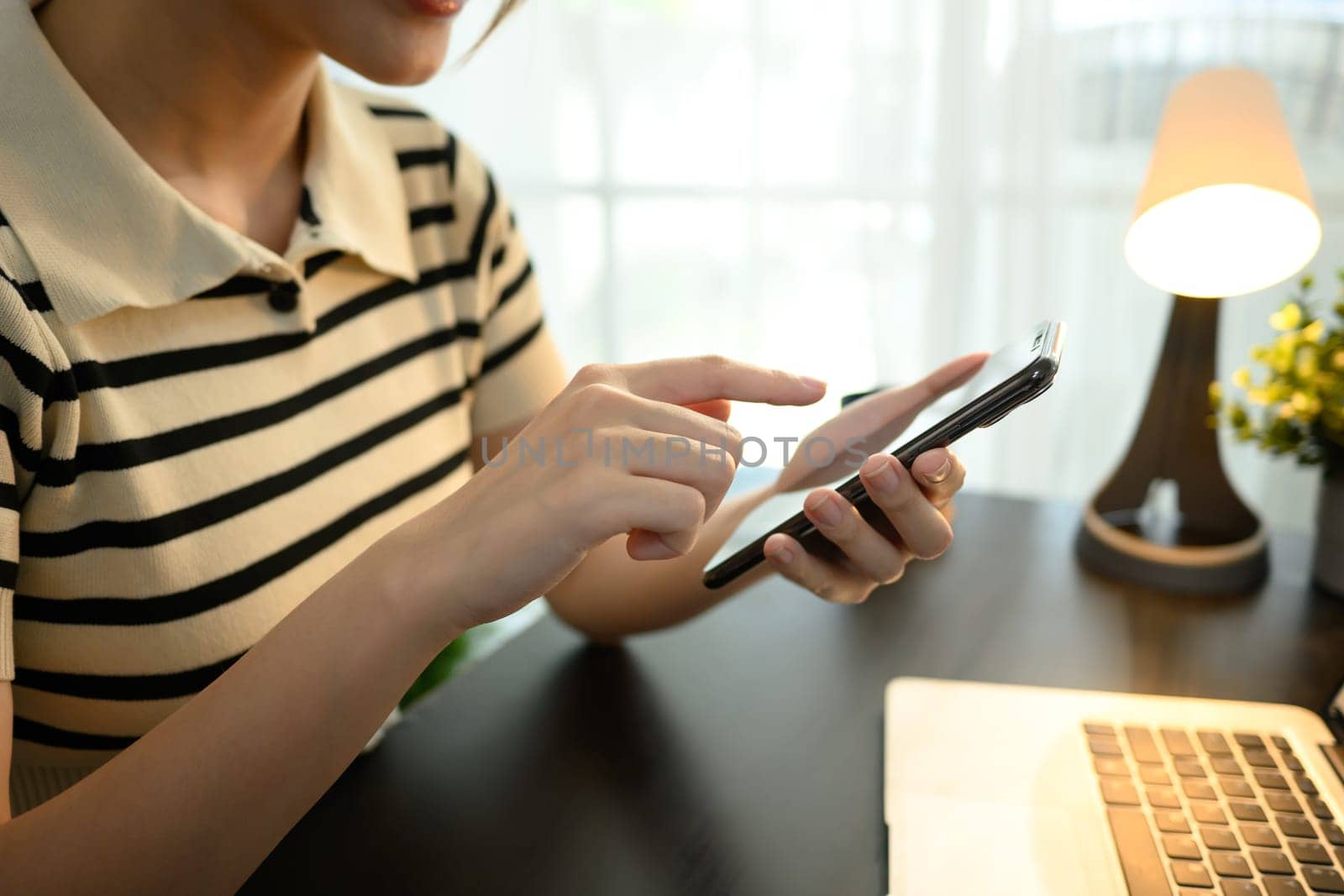 Cropped shot young woman using smartphone while working at home office desk with laptop.