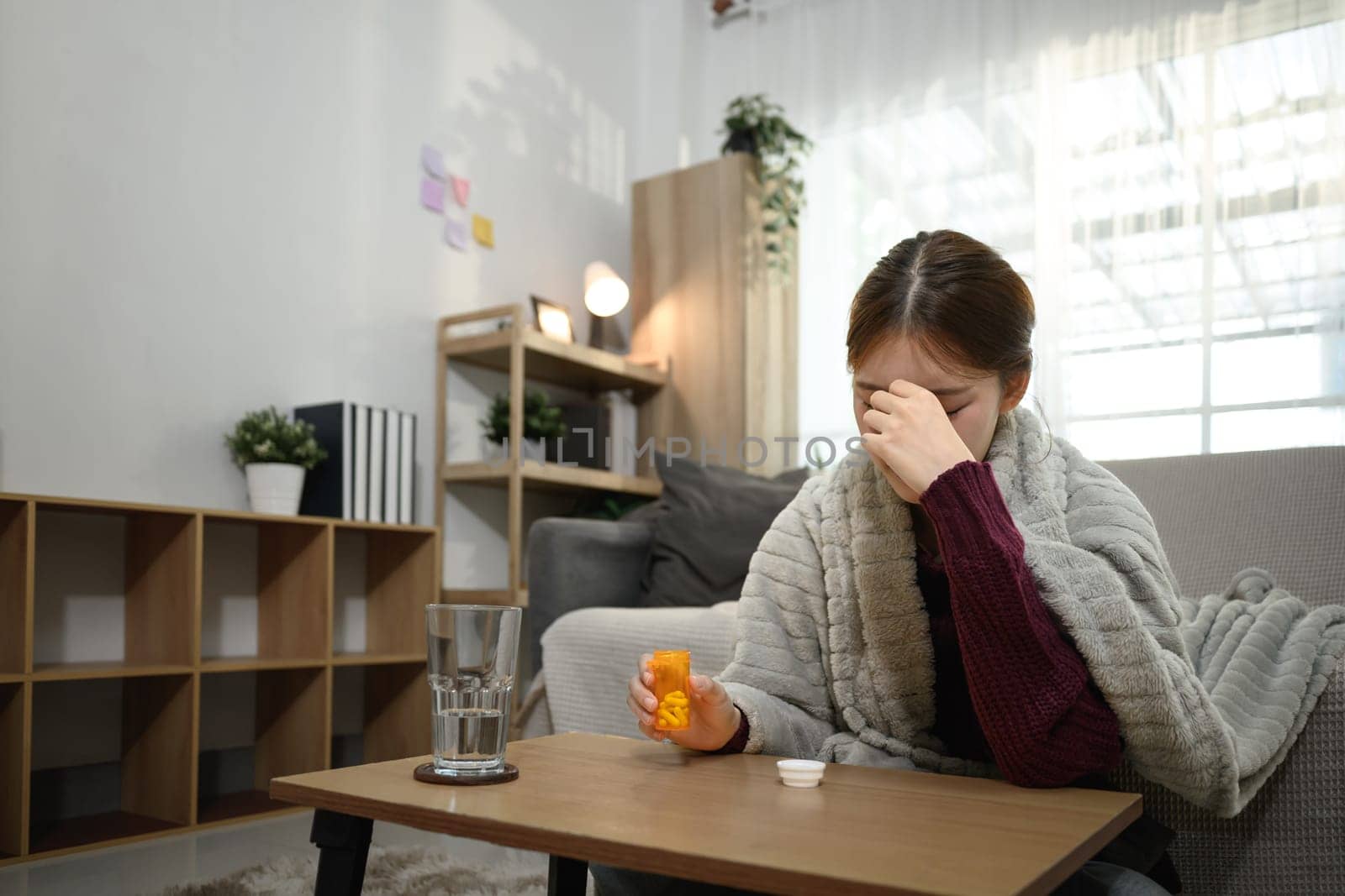 Sick young woman wrapped in blanket holding a pill bottle while experiencing discomfort or headache.