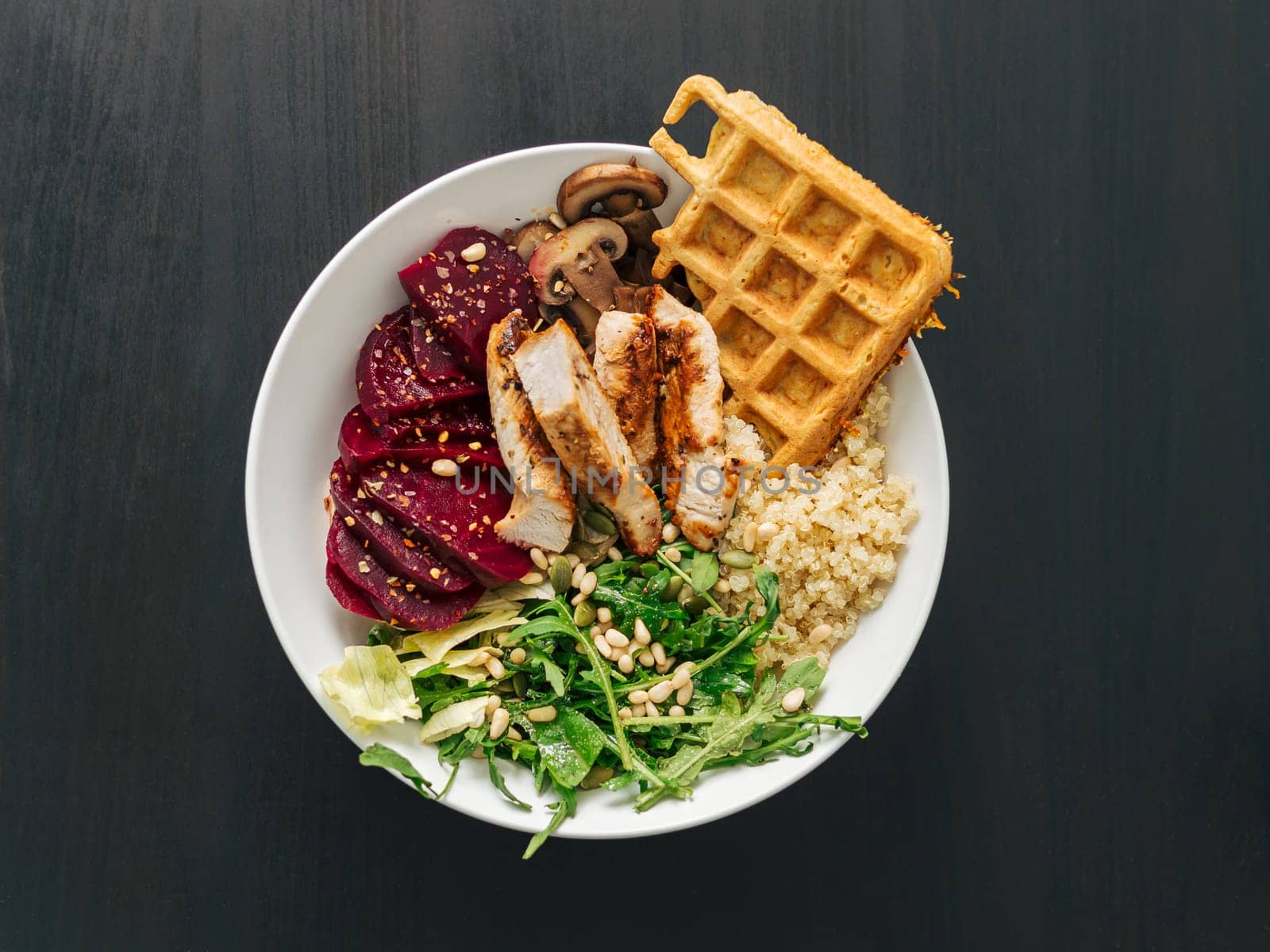Healthy Food concept. Bowl with chicken, mushrooms, beetroot, quinoa, green rucola, decorated pine nuts and carrot waffle. Top view of gourmet bowl. Balanced gourmet dish on black wooden background