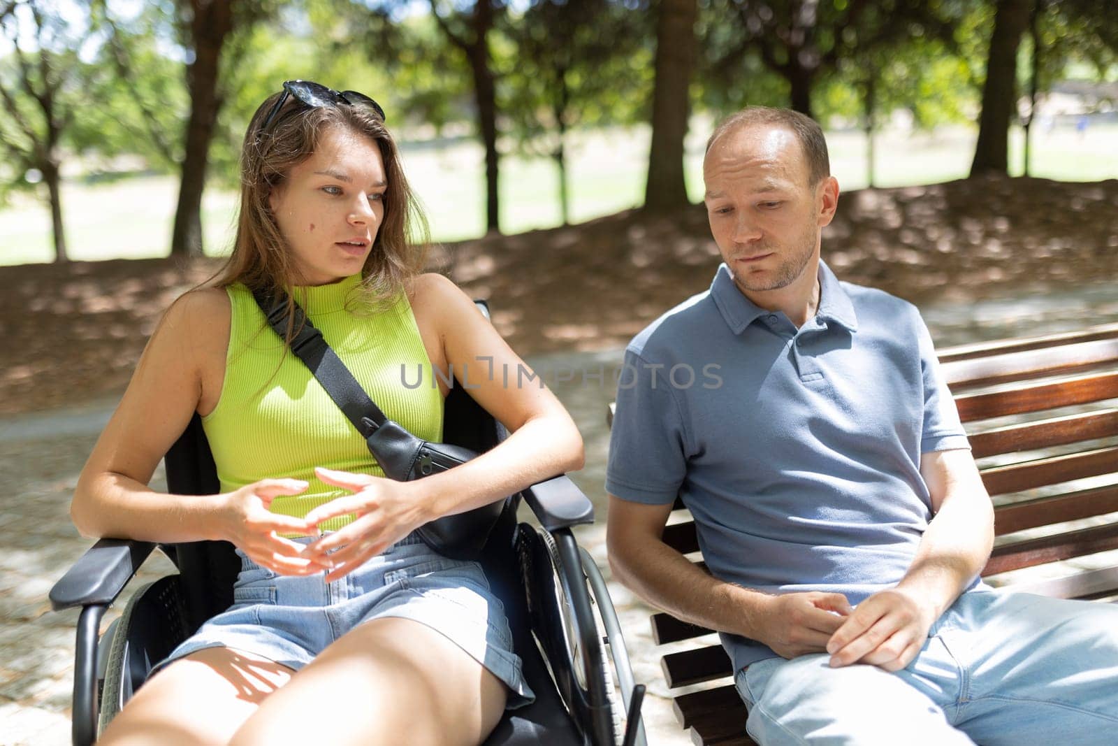 Serious young woman in wheelchair explaining something to man sitting next to her on park bench