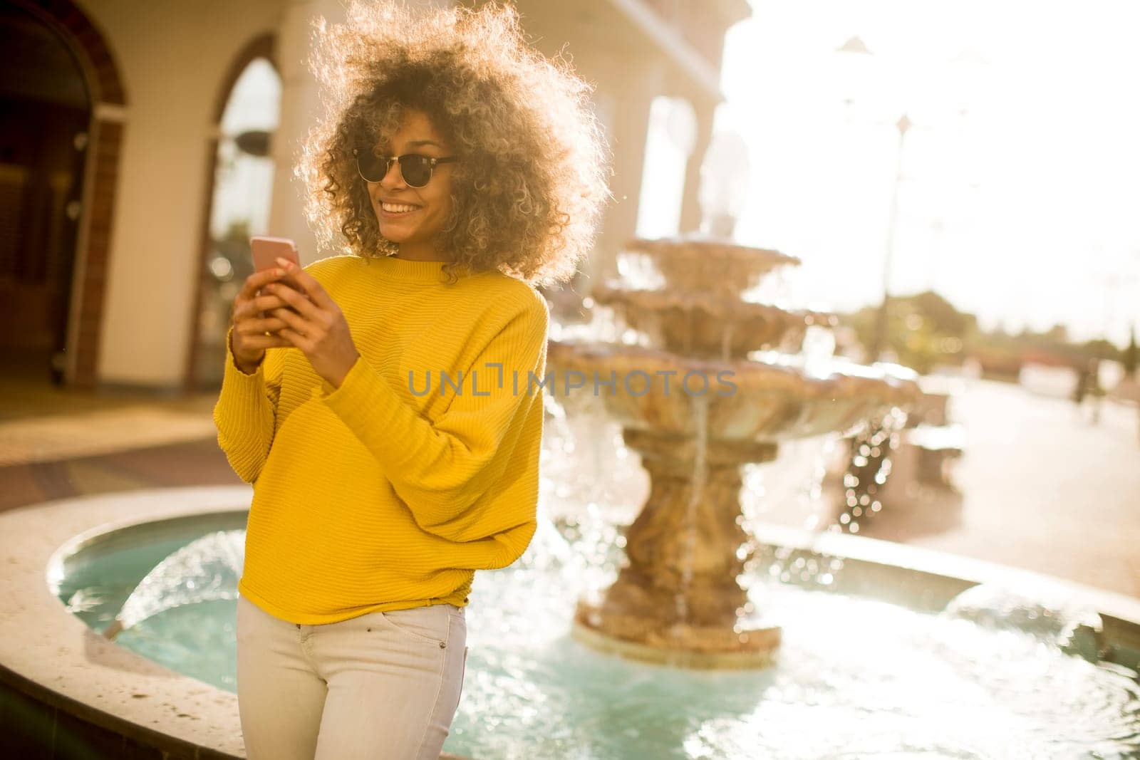 Attractive young woman with curly hair using her touch screen mobile cell phone by the fountain