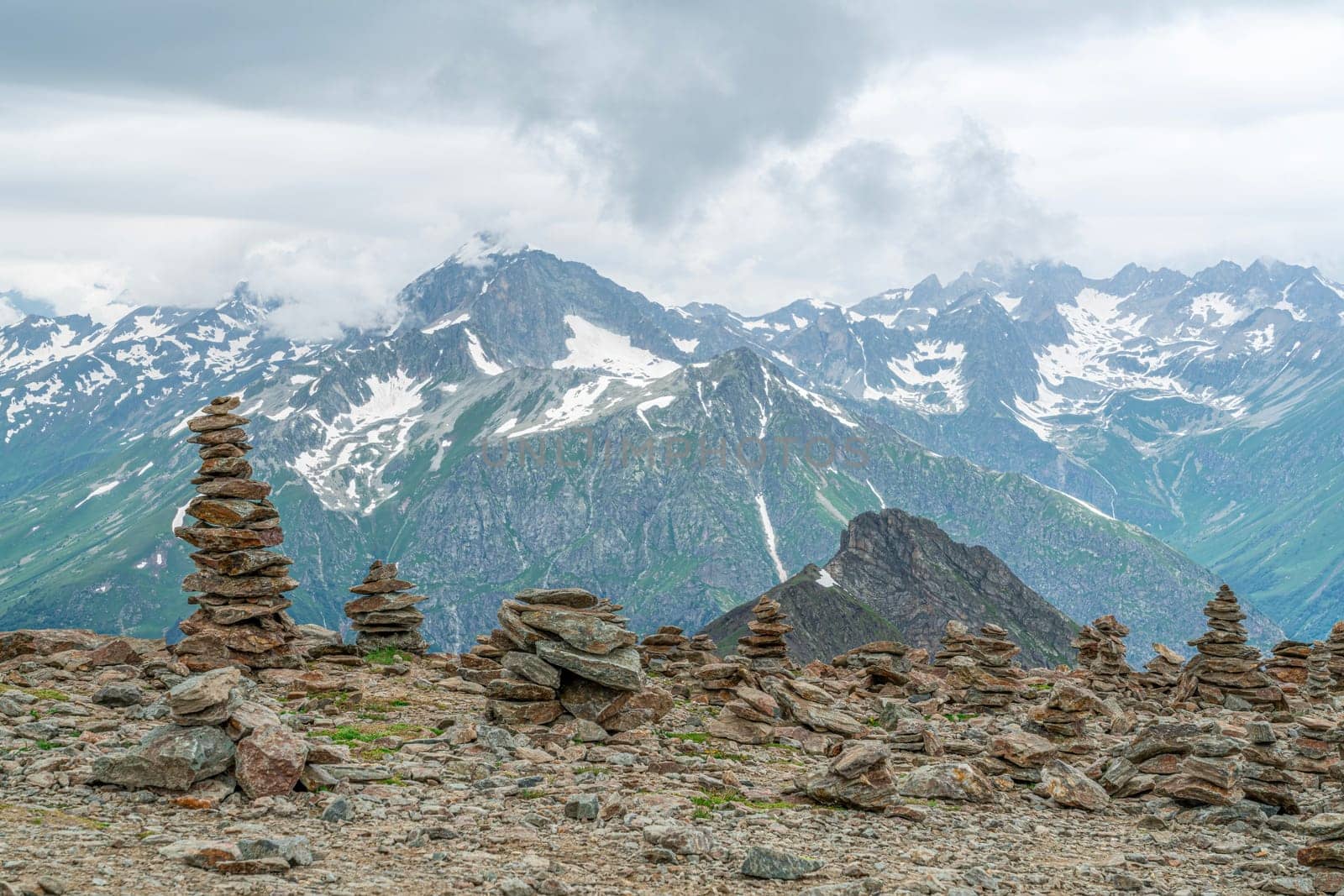A breathtaking view of towering mountain peaks, adorned with patches of snow, contrasts with carefully stacked stone cairns on the rocky terrain. The clouds loom above, enhancing the atmosphere.