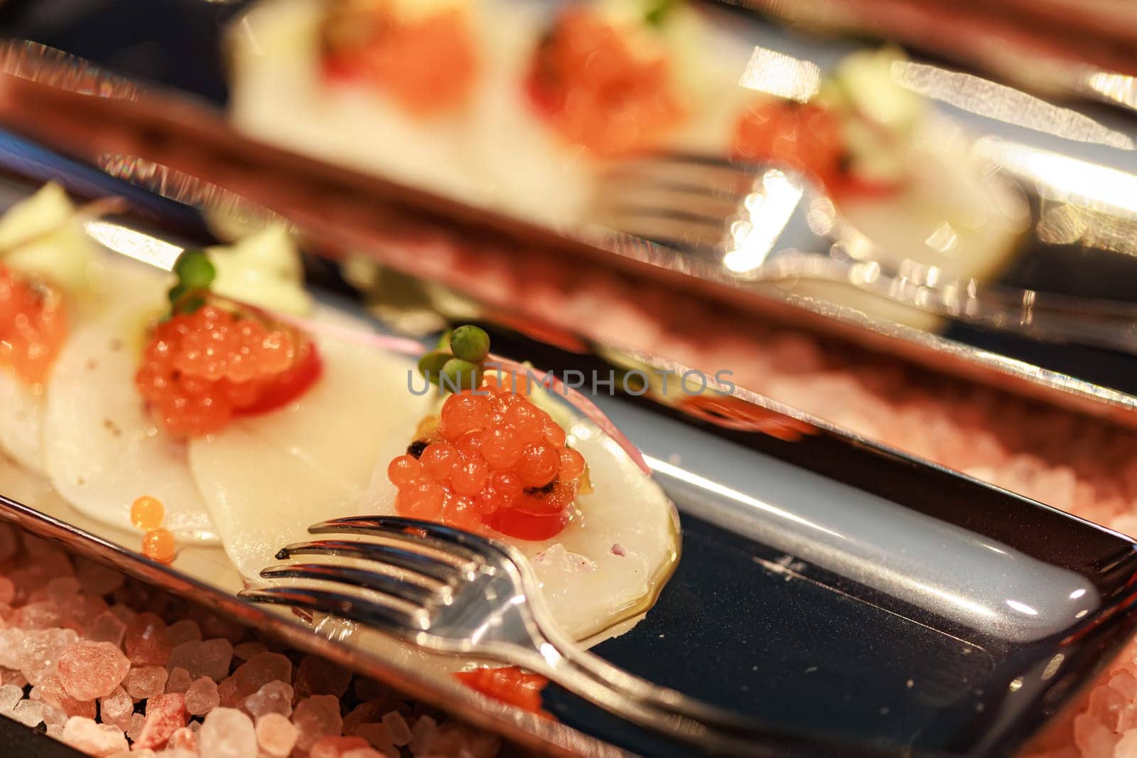 Elegant seafood appetizer featuring thinly sliced scallops, garnished with vibrant salmon roe, cherry tomatoes, microgreens, and a delicate sauce, artistically presented on sleek plates.