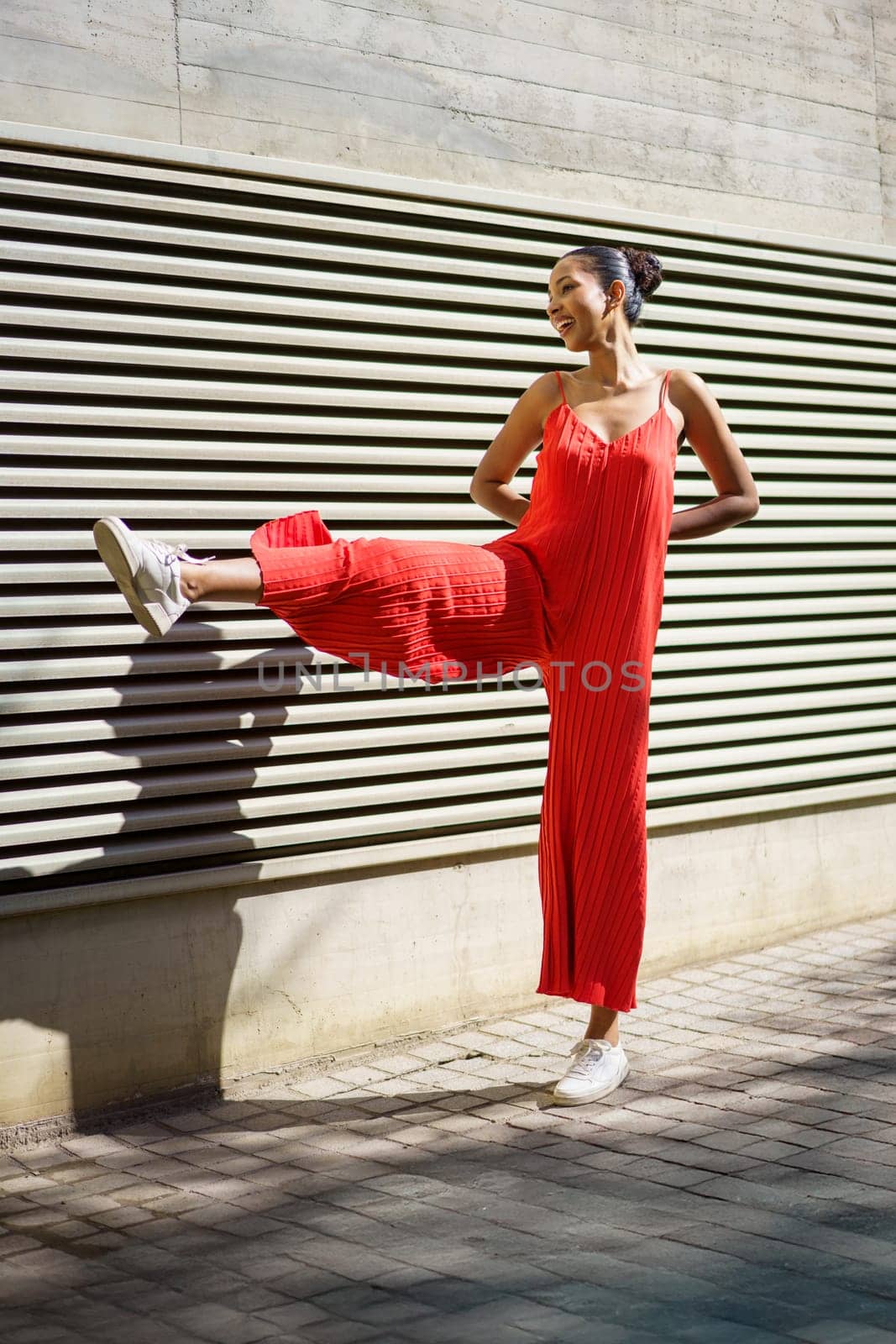 A cheerful and vibrant young woman showcases her bold red attire while playfully posing against a beautifully textured wall, perfectly combining elements of fashion and fun in her dynamic expression