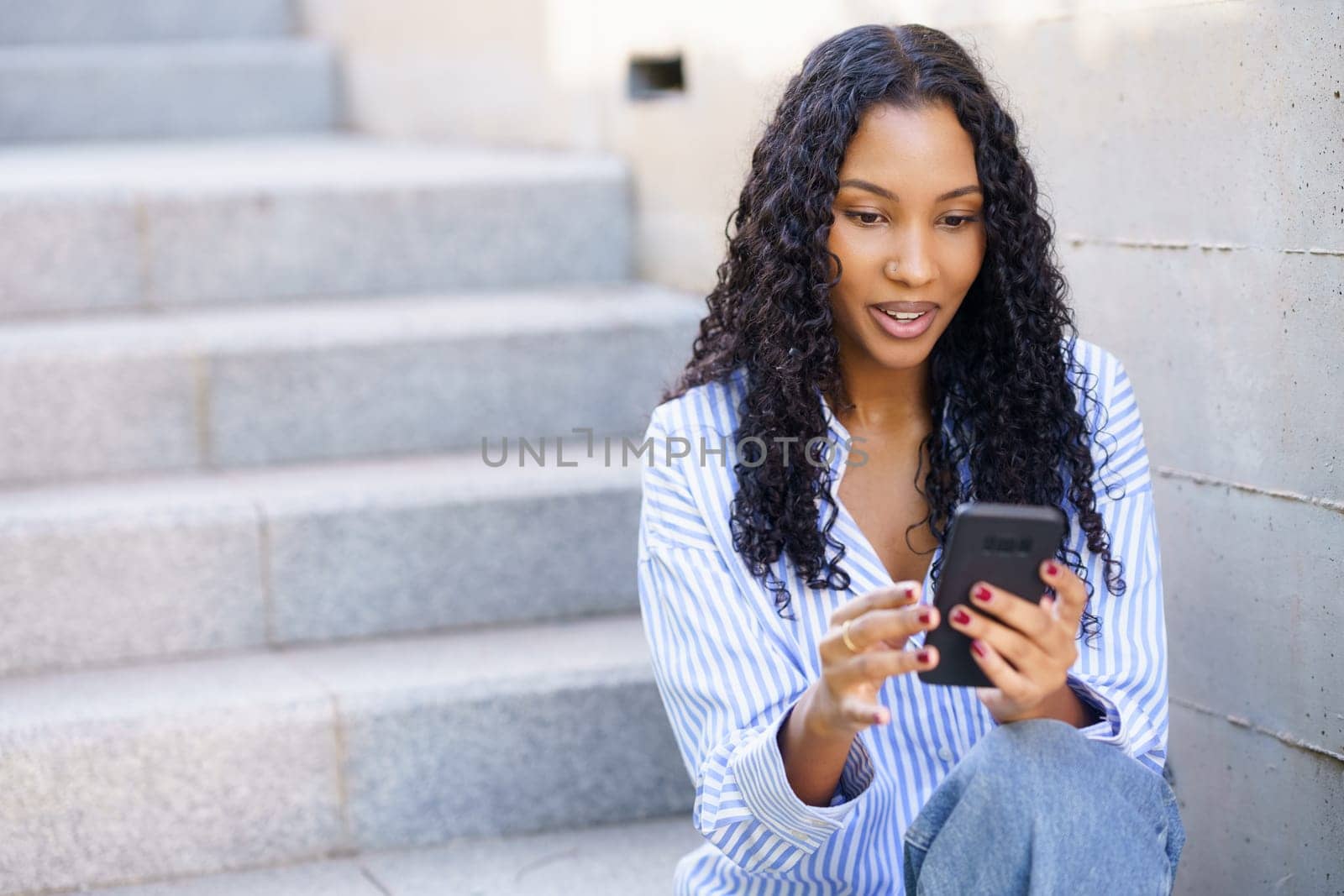 In a vibrant urban setting, a young woman with bouncy curly hair sits on outdoor steps, fully engaged with her smartphone, symbolizing the modern lifestyle many aspire to in their busy lives