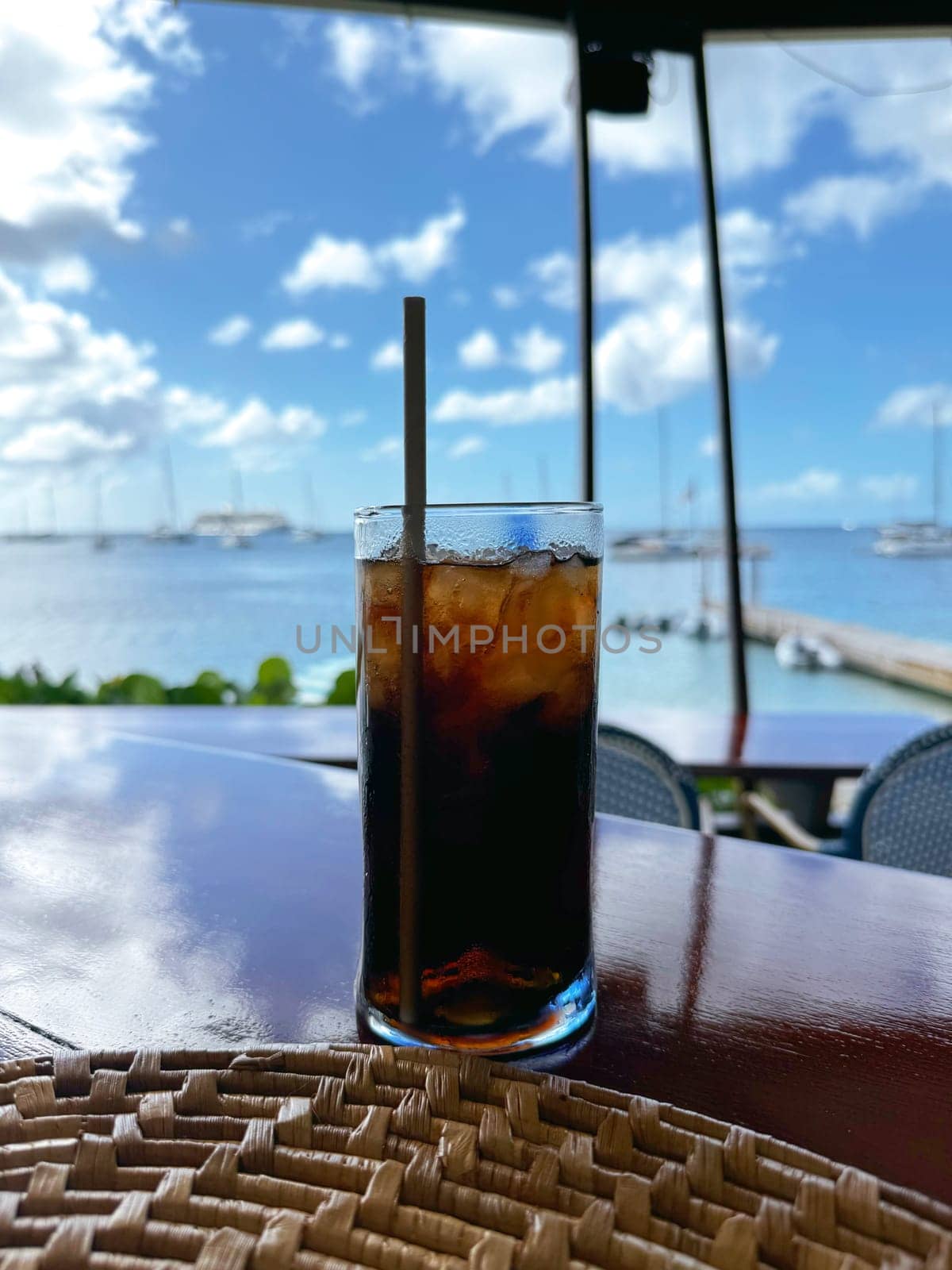 Refreshing glass of iced cola with a straw on a bar counter, overlooking a tropical marina filled with boats and sunshine.