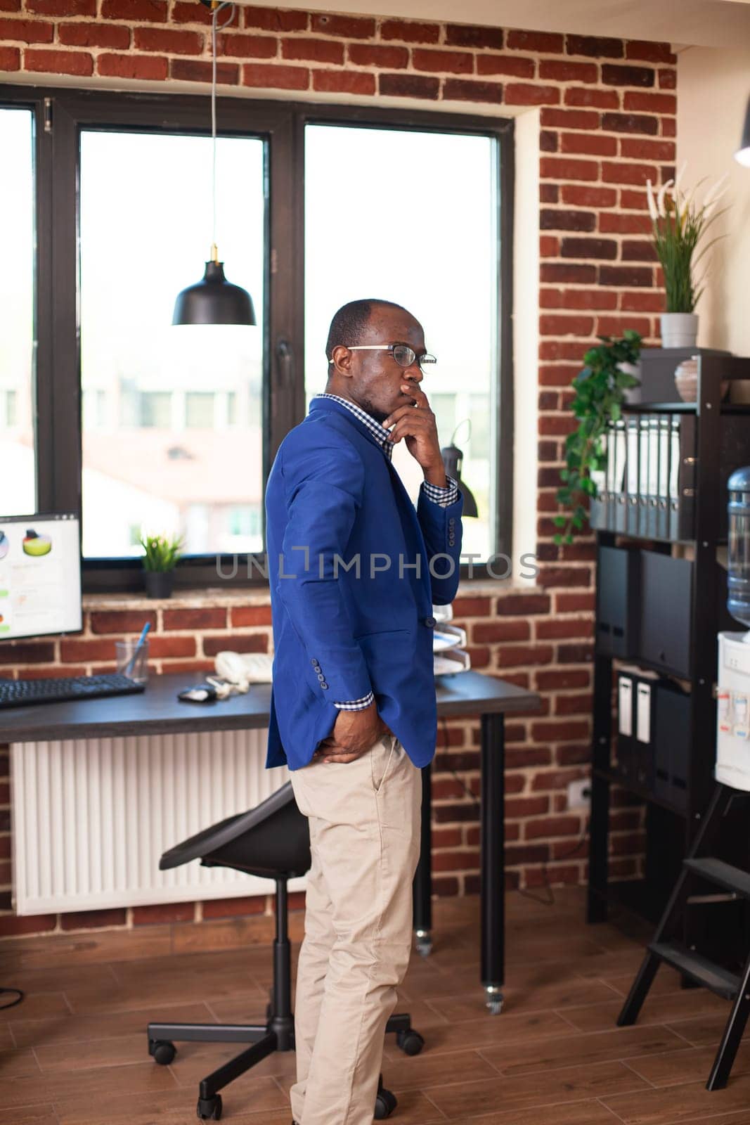 Black man with hand over mouth, standing in empty room and reflecting on startup business concepts. Male entrepreneur in brick wall workspace, contemplating ideas and observing office decor.
