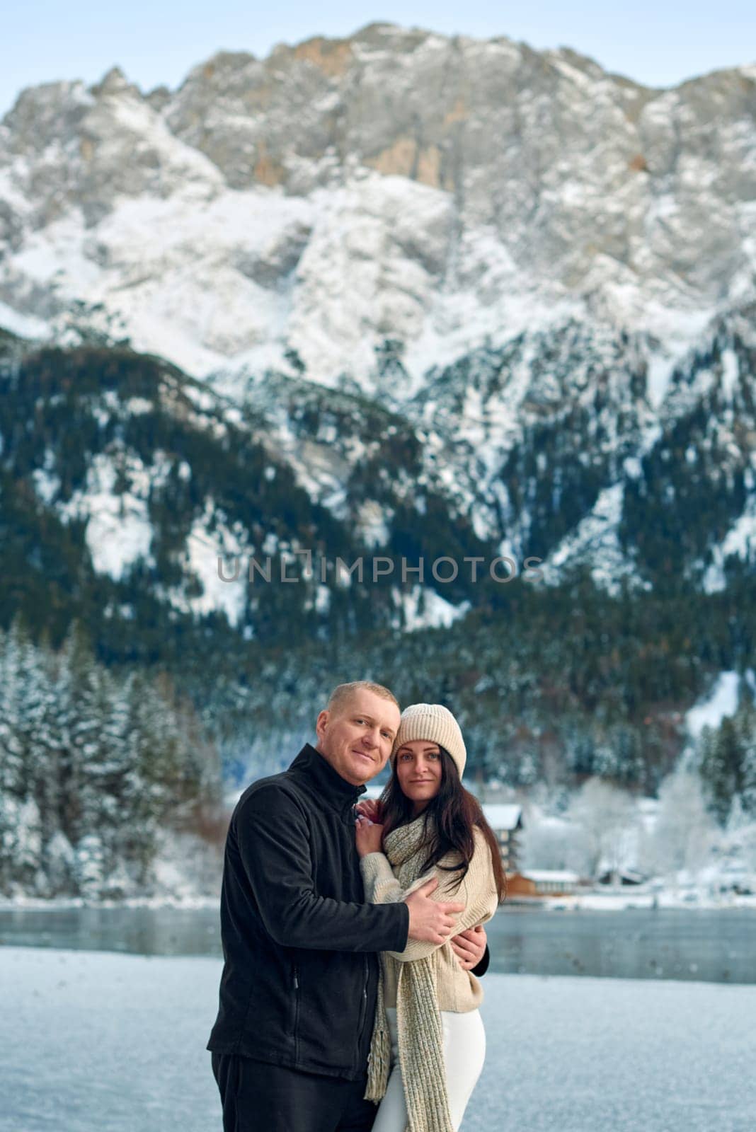 Romantic Winter Getaway Couple on Frozen Alpine Lake with Snowy Mountains and Rustic Cabin. Serene Outdoor Portrait of Man in Black Fleece and Woman in Cream Sweater Enjoying Picturesque Landscape - This high-resolution winter photograph captures a romantic couple enjoying a serene moment on a frozen alpine lake. The man, with short light brown hair, wears a black fleece jacket, dark pants, and white sneakers, while the woman, with long dark brown hair, is dressed in a beige knitted beanie, cream-colored chunky knit sweater, white leggings, and a long cream scarf. Their relaxed and content expressions convey a sense of peace and togetherness against the breathtaking winter backdrop. The background features snow-covered mountains densely forested with evergreen trees, their branches laden with snow. A rustic wooden cabin with a snow-covered roof nestles among the trees, adding a cozy element to the scene. The partially frozen lake reflects the surrounding landscape, creating a mirror-like effect that enhances the tranquil atmosphere. This image perfectly encapsulates the beauty of winter outdoor activities, romantic getaways, and the serene majesty of alpine landscapes, making it ideal for travel, lifestyle, and seasonal content.
