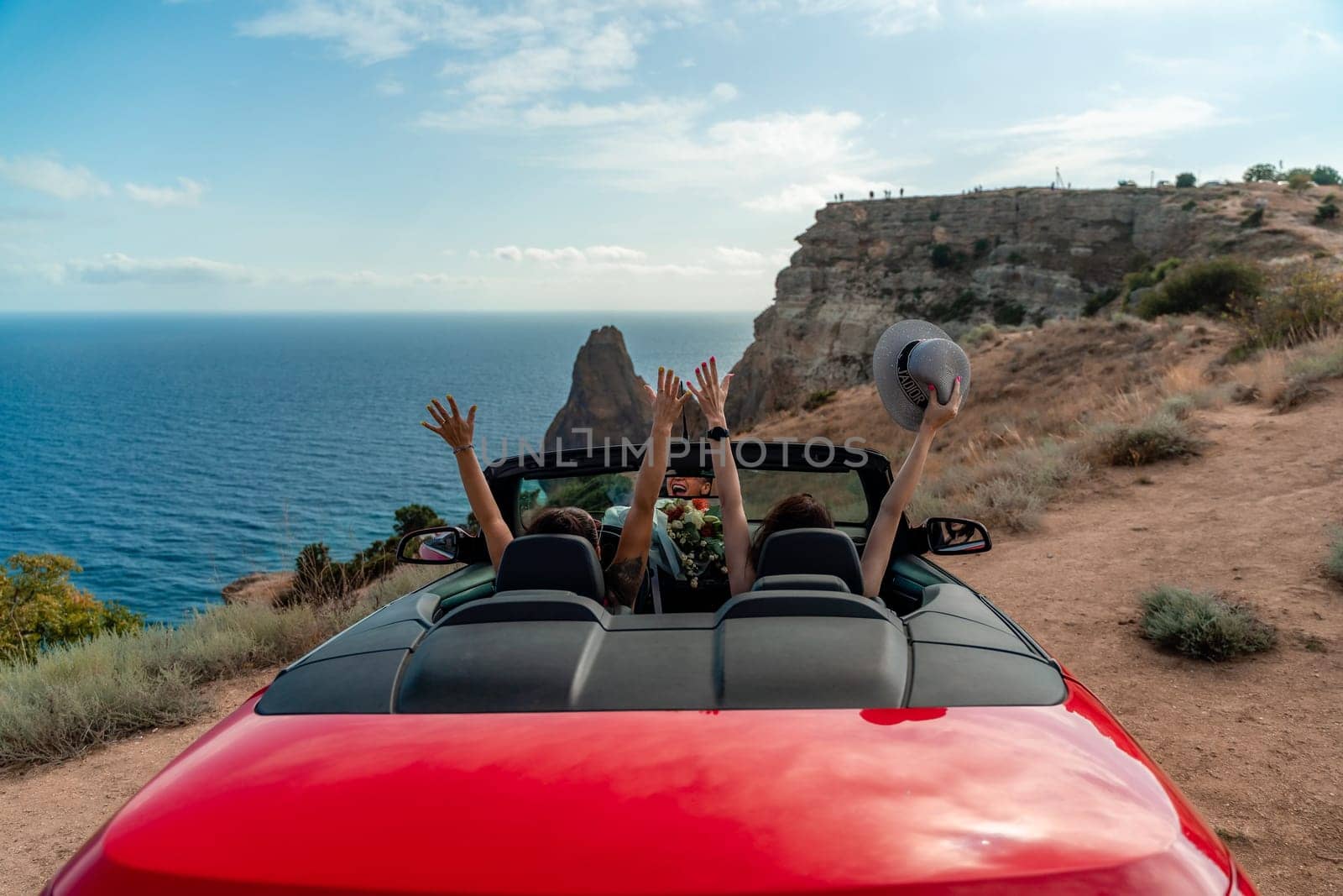 A red convertible car with two people in it, one of whom is waving. The car is driving on a road near the ocean