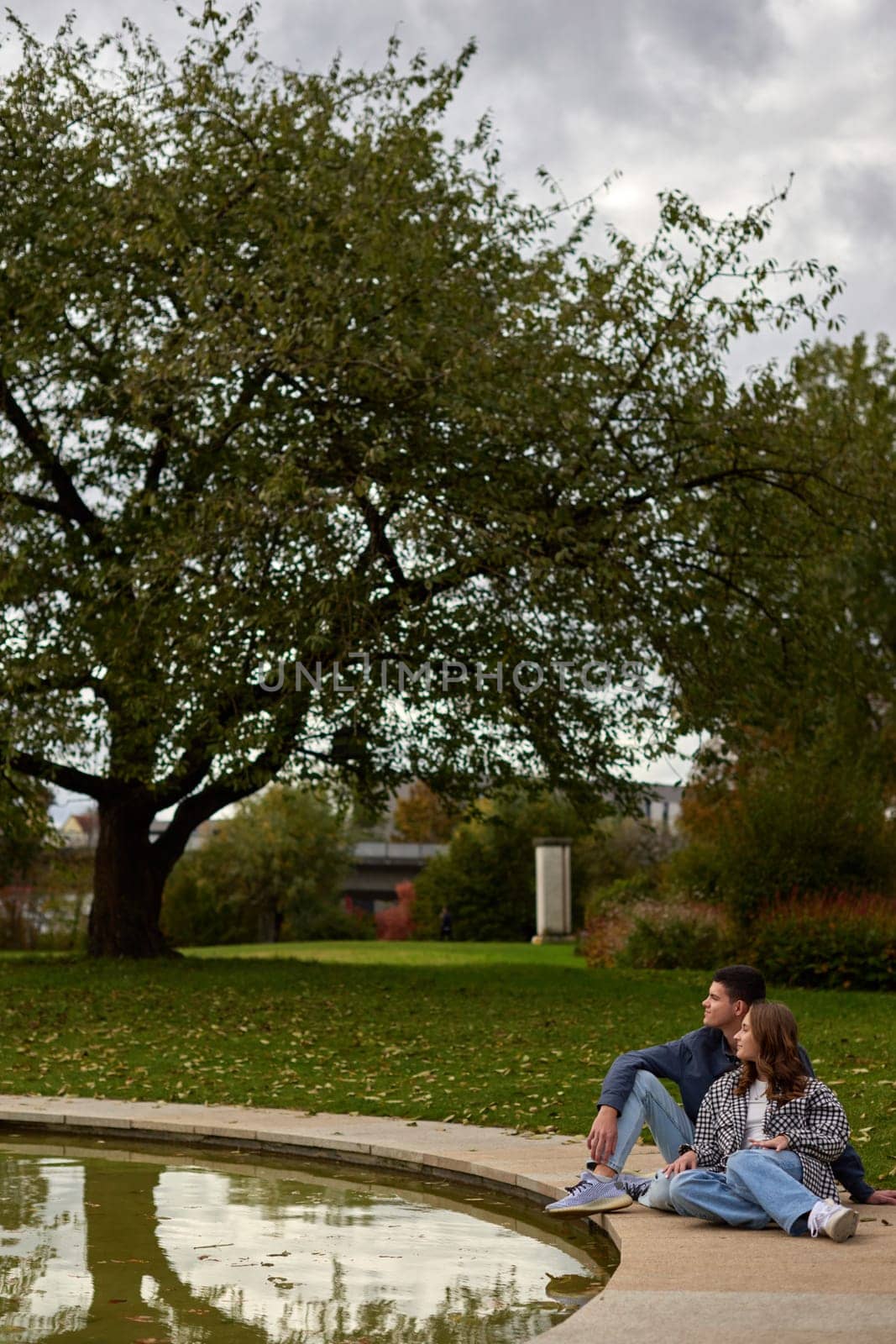 This picturesque scene captures two individuals sitting on the grass by the edge of a reflective pond in a serene park or garden. The couple is leaning against each other, enjoying a moment of relaxation surrounded by the vibrant colors of autumn. The background features trees adorned with red and orange leaves, and a building covered in lush green ivy. The reflection of the couple in the pond adds to the tranquil atmosphere, creating a beautiful and peaceful setting. Autumn Serenity: Couple Relaxing by a Reflective Pond in an Ivy-Covered Garden with Vibrant Foliage