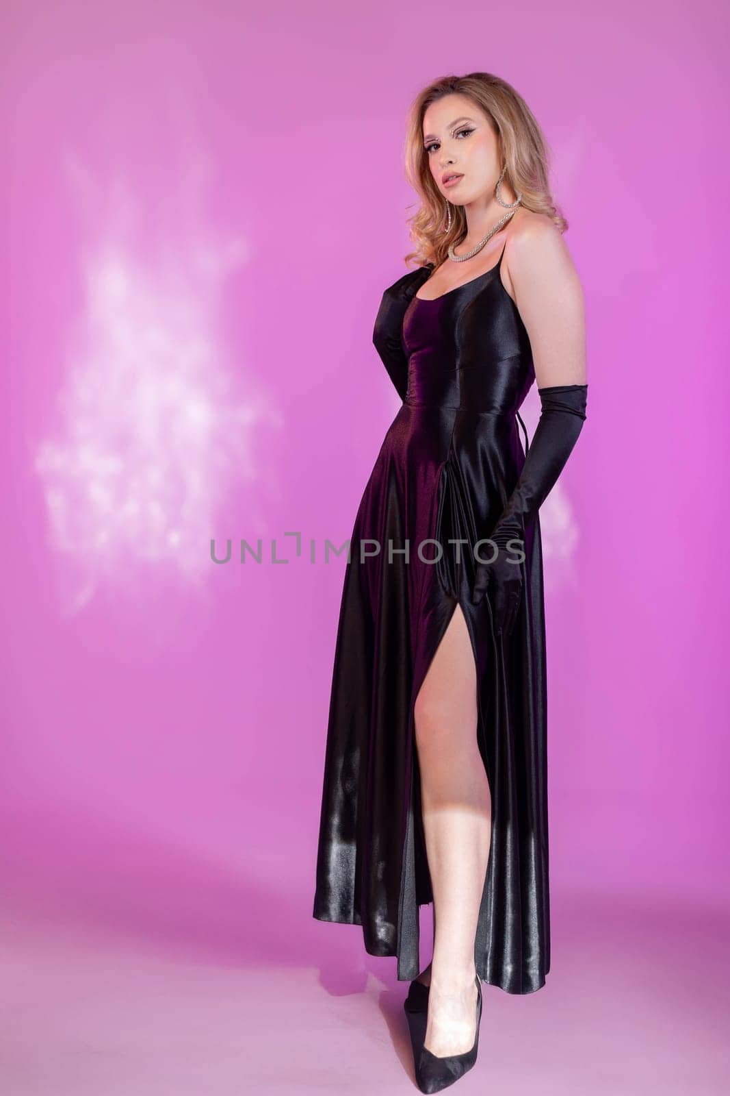 A stylish woman poses elegantly while wearing a long, flowing black dress against a vibrant pink backdrop
