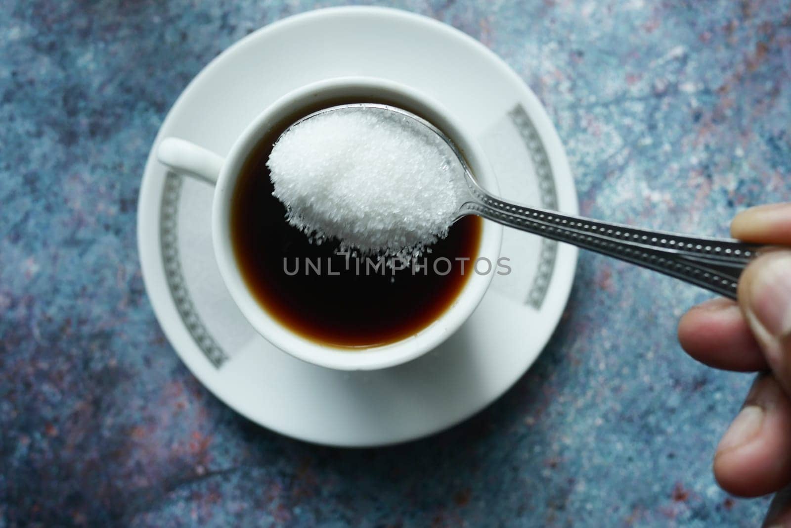 A hand holds a spoonful of sugar over a steaming cup of coffee.